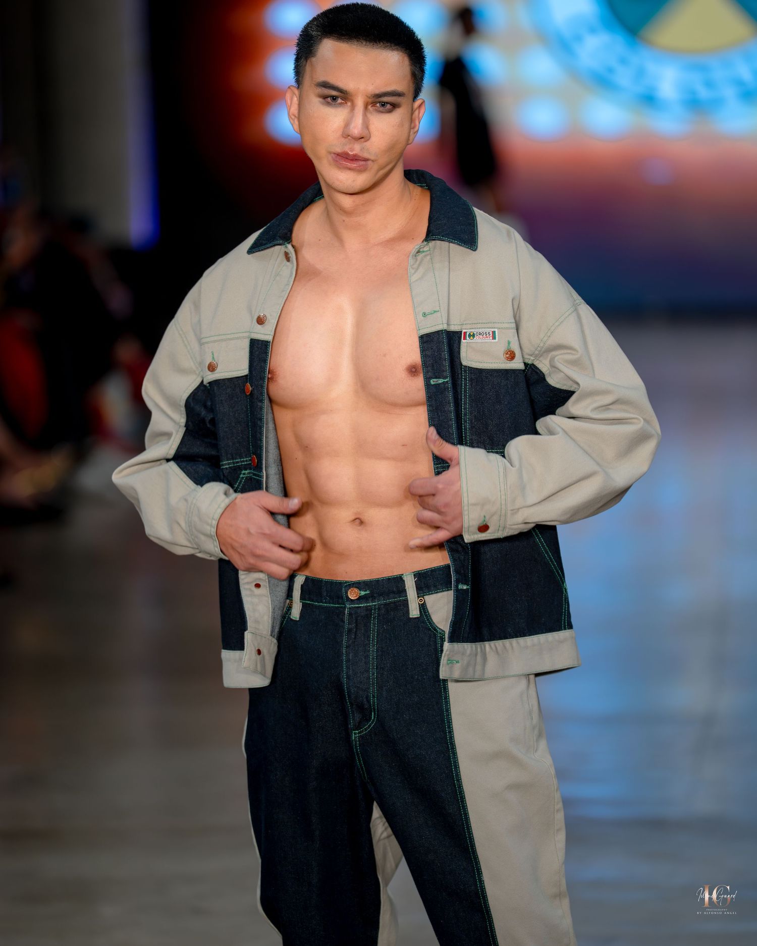 Model walks runway wearing denim jeans and light jacket showing bare chest during fashion show with blue lighting.