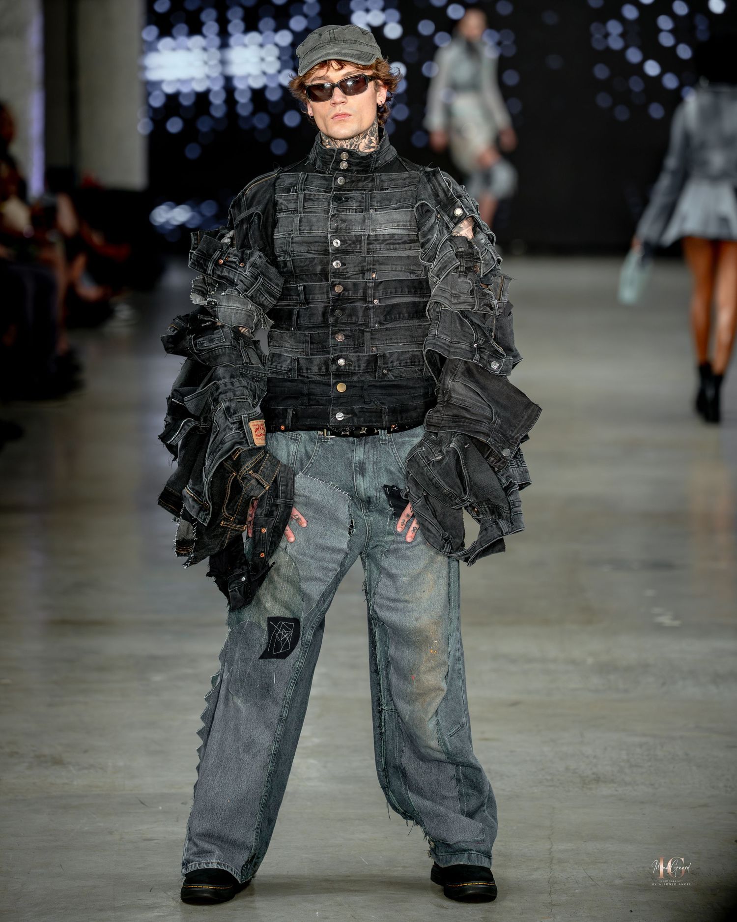 A model walks the runway wearing tactical military-inspired gear with a black vest and distressed jeans at a fashion show.