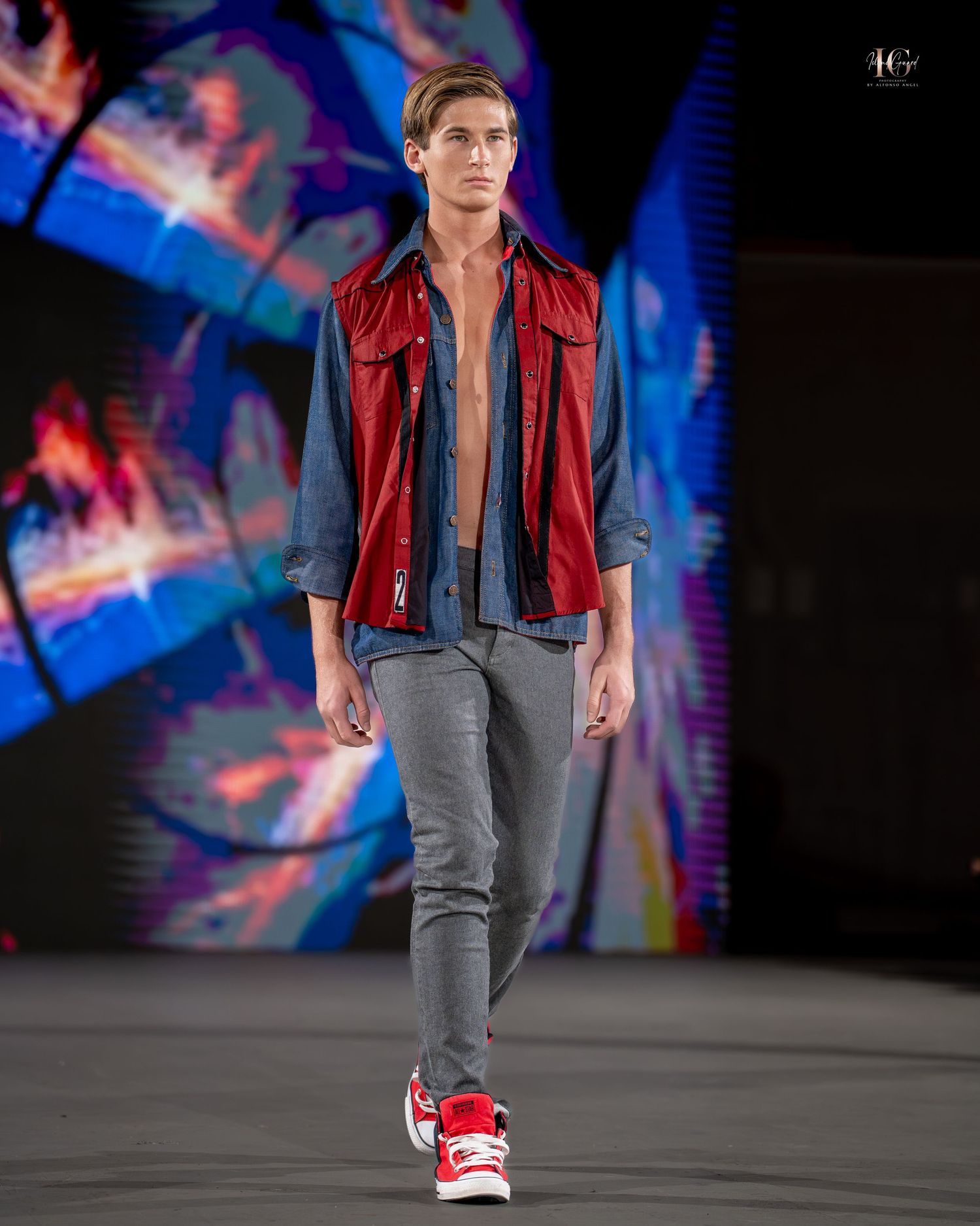 Model walks runway wearing red puffy vest over grey outfit with red sneakers at fashion show.