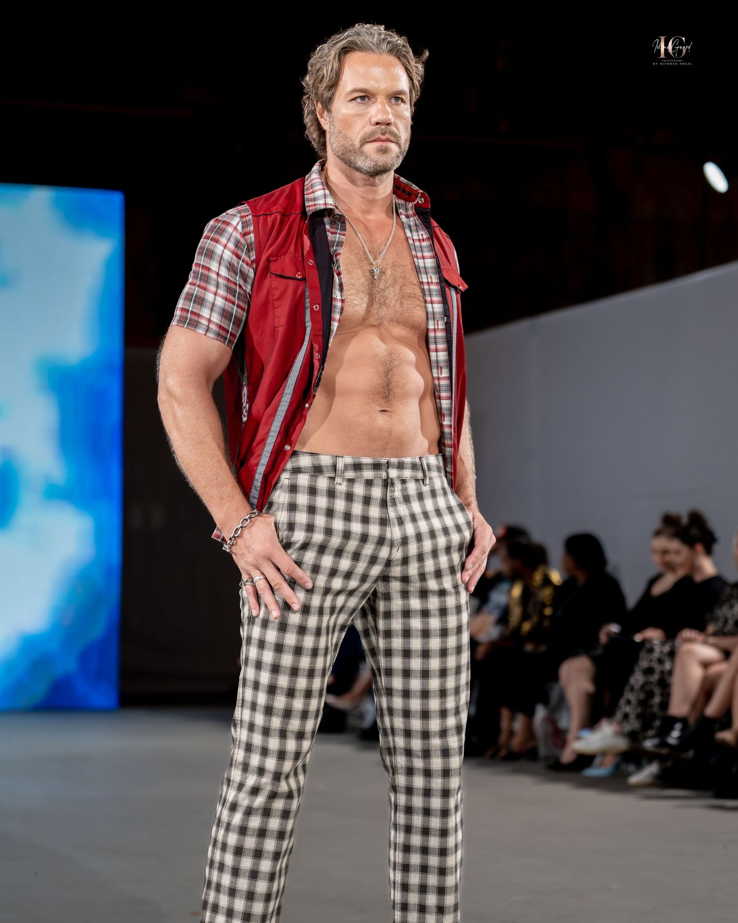 Model walks runway wearing red vest and black and white checkered pants during fashion show.