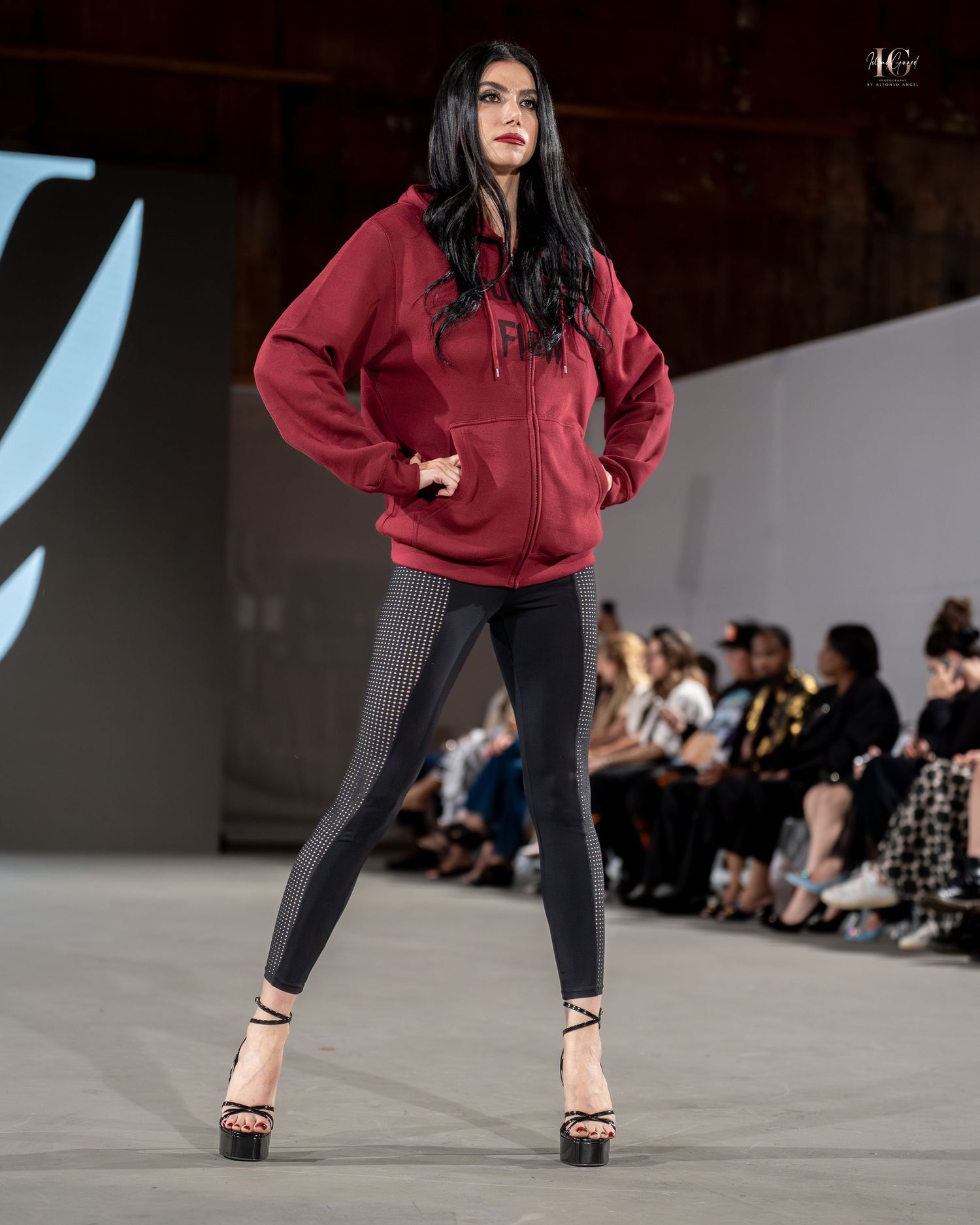Model walks runway in burgundy hoodie and black leggings at fashion show, striking dynamic poses on concrete floor.