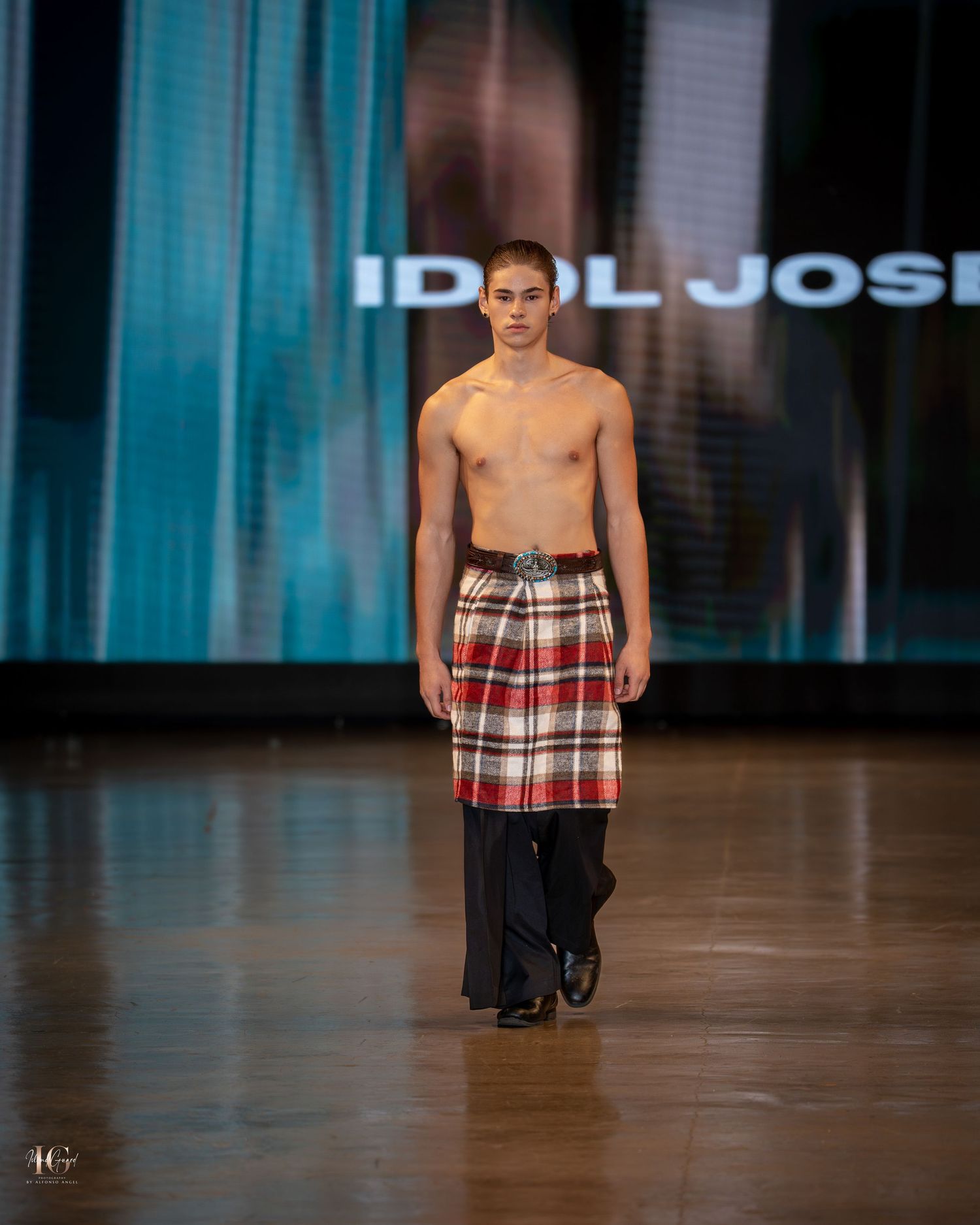 A model walks the runway in a plaid kilt and black top during a fashion show on a wooden floor.