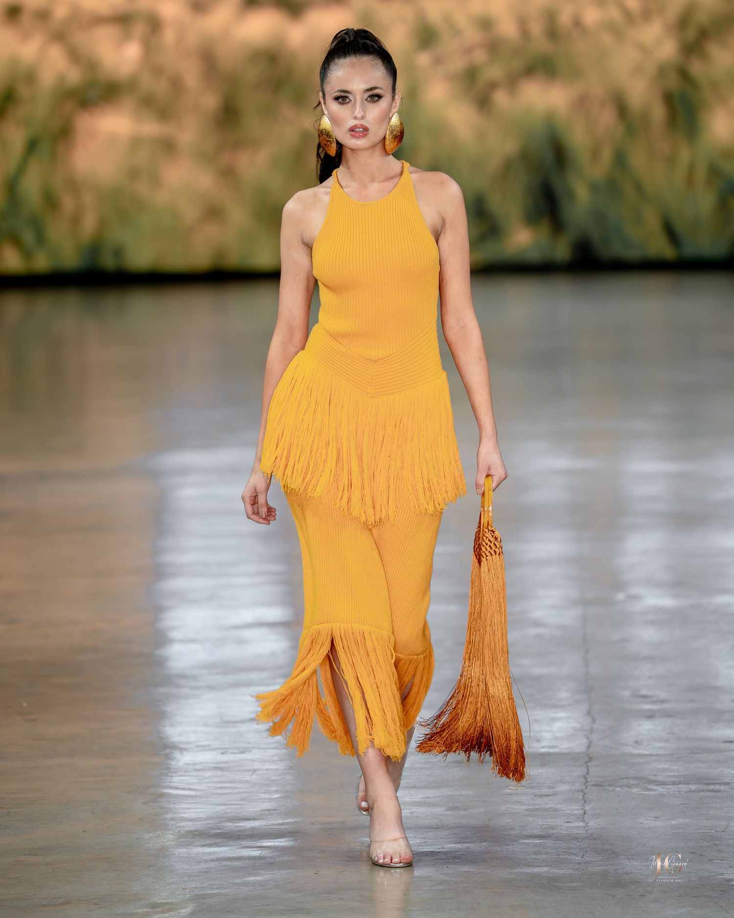 Model walks runway in flowing marigold yellow dress with tiered ruffles and fringe details at outdoor fashion show.