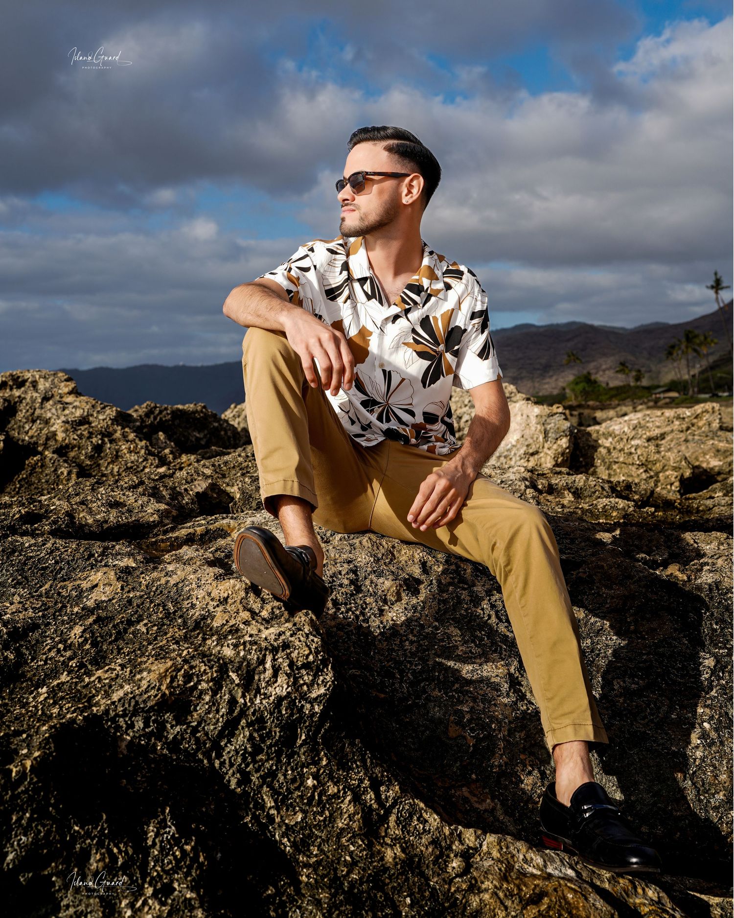 Person wearing sunglasses and floral print shirt sits on coastal volcanic rocks with mountains in background.