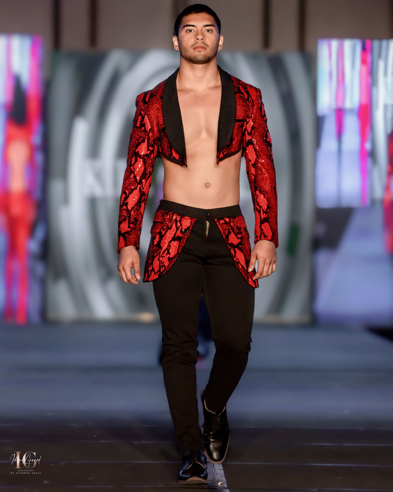 Fashion model walks runway in red lace blazer with black pants against dramatic lighting backdrop.