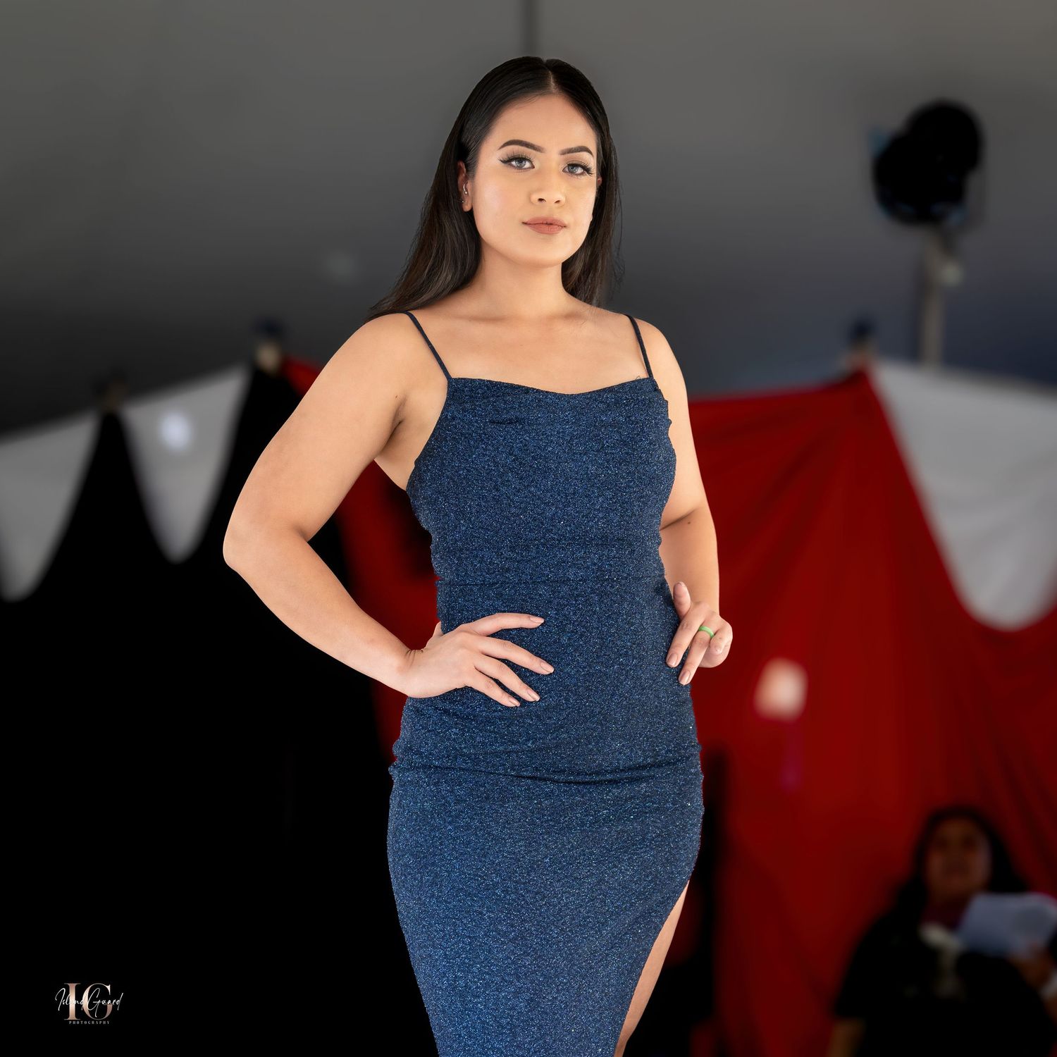 Model in fitted blue denim dress poses against dark studio backdrop with lighting equipment visible.