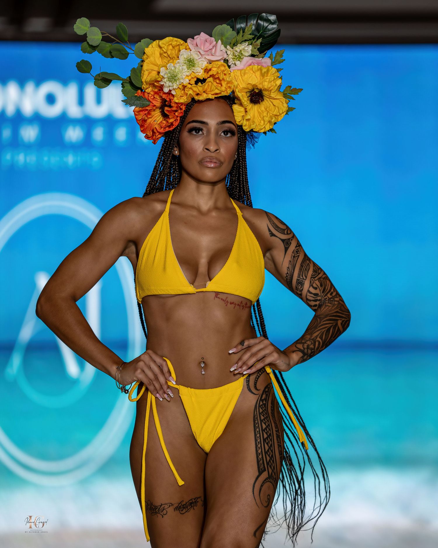 Model walks runway in yellow bikini with floral crown and tribal tattoos at Honolulu Swim Week fashion show.