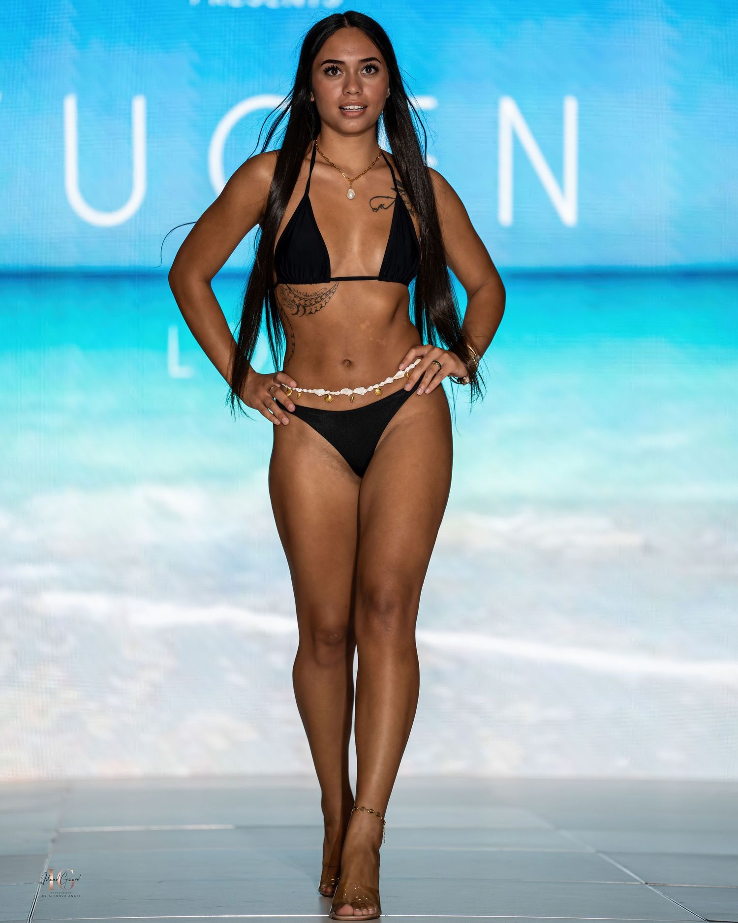 Model showcases black bikini with gold waist chain at beachwear fashion show.