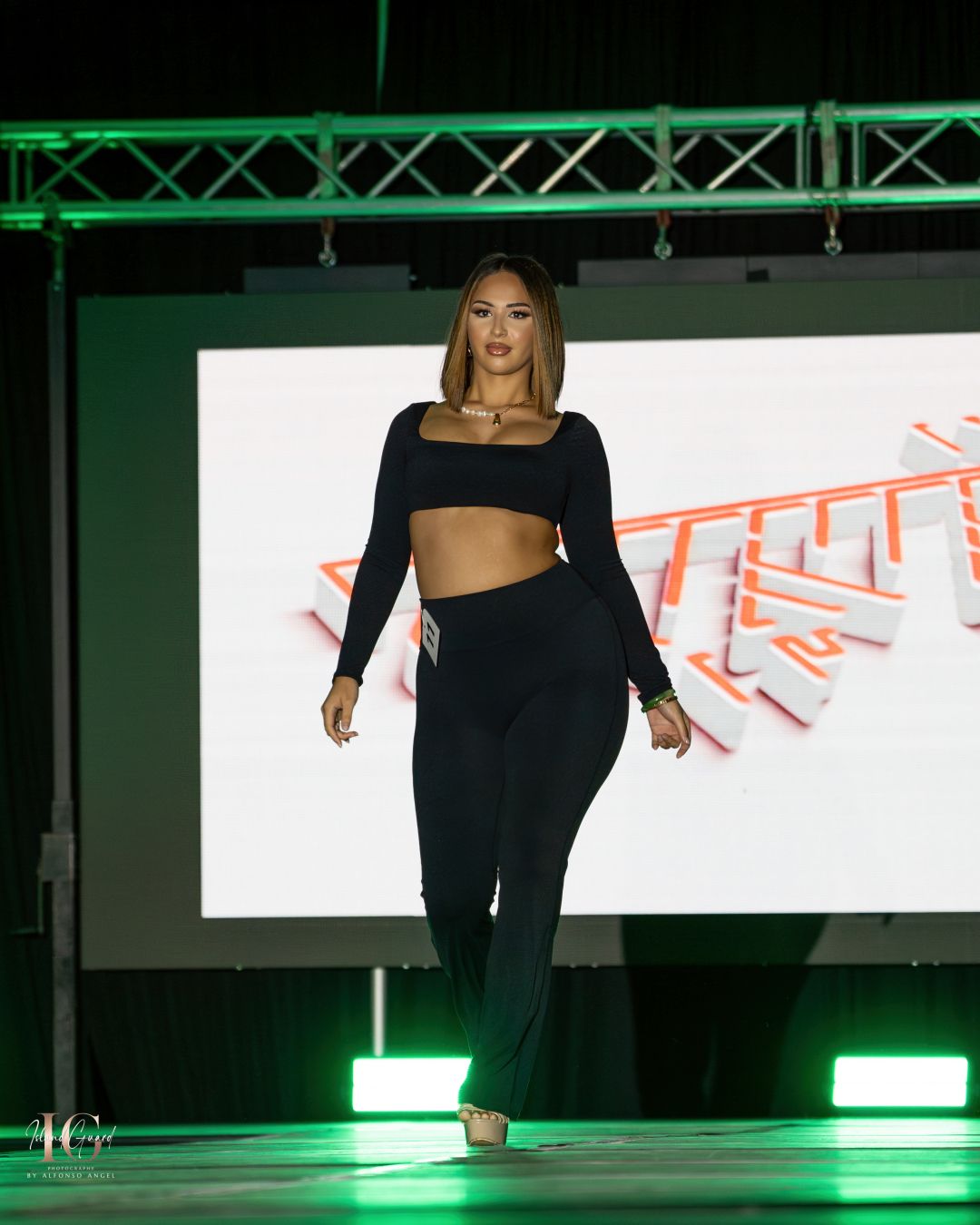 Fashion model showcases black crop top and fitted pants ensemble on illuminated runway with stage lighting.