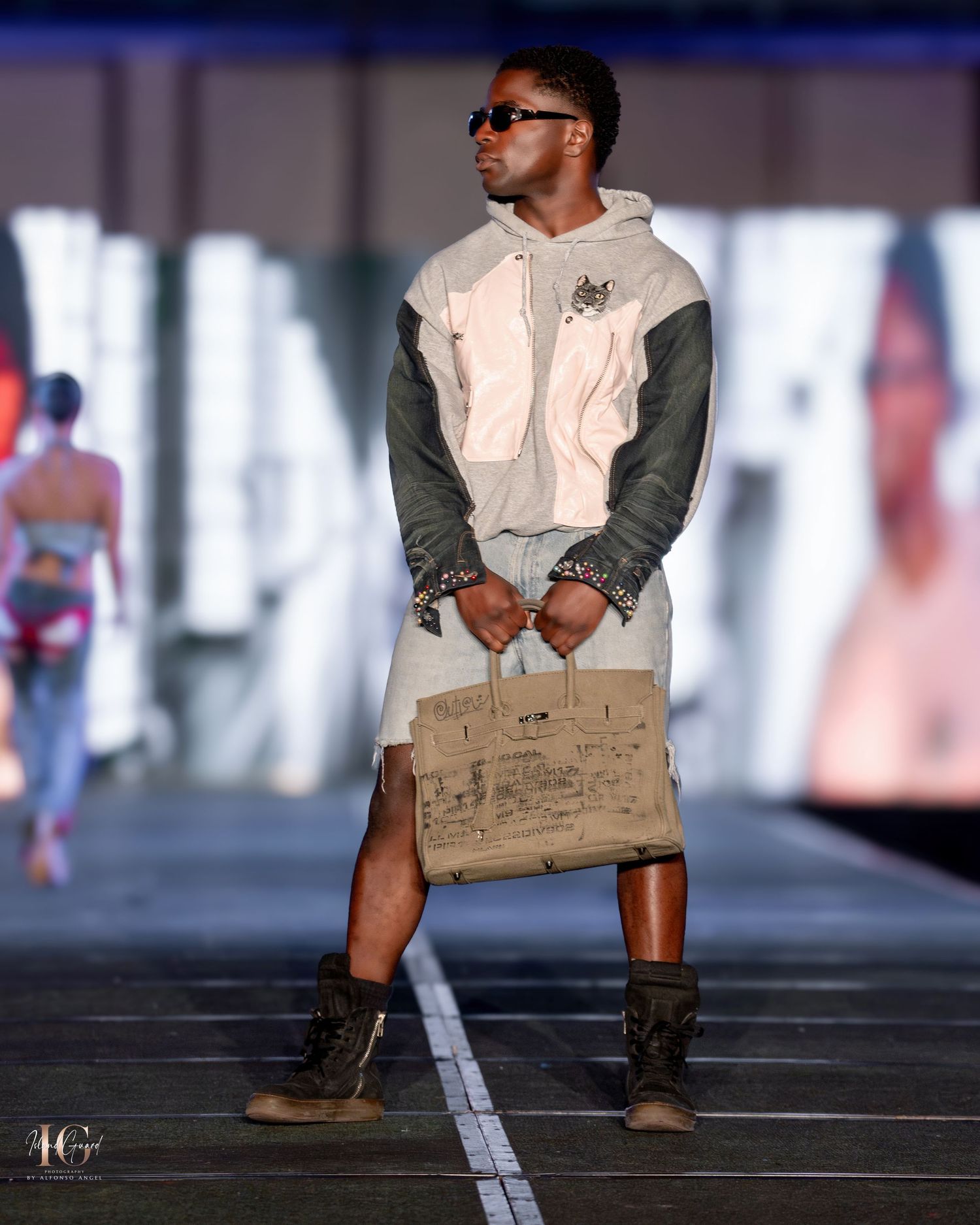 Model in two-tone hoodie and shorts carries brown paper bag accessory on fashion runway.