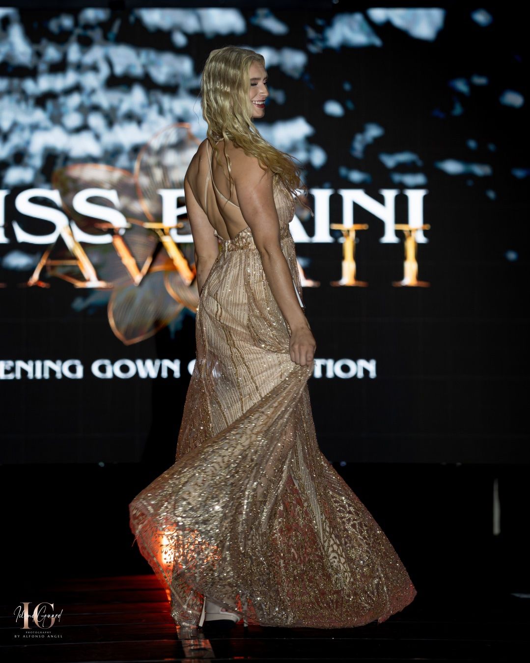 Model walks runway wearing shimmering gold evening gown with flowing skirt at glamorous fashion show event.