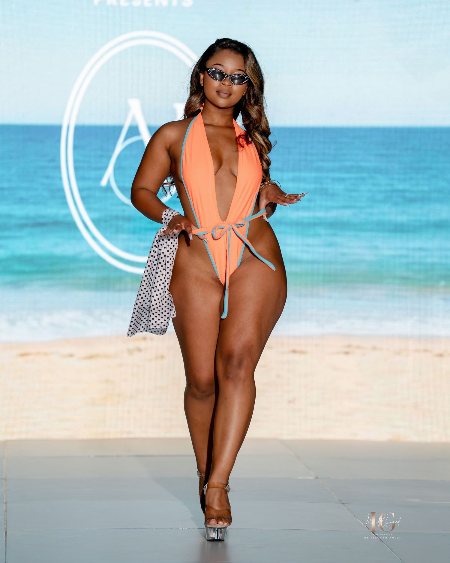 Model poses on sandy beach wearing orange swimsuit and carrying polka dot bag against turquoise ocean backdrop.