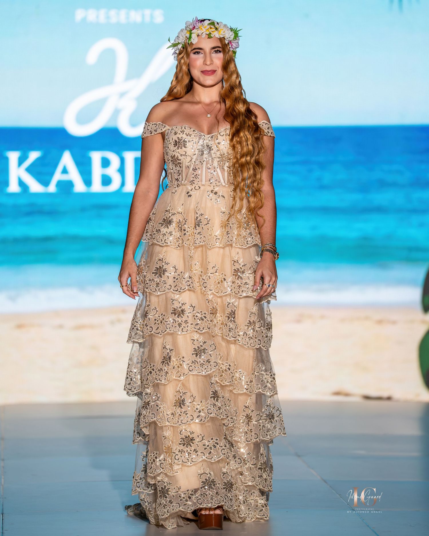 A model wearing a floral beige tiered lace gown with a flower crown walks the runway at a beach-themed fashion show.