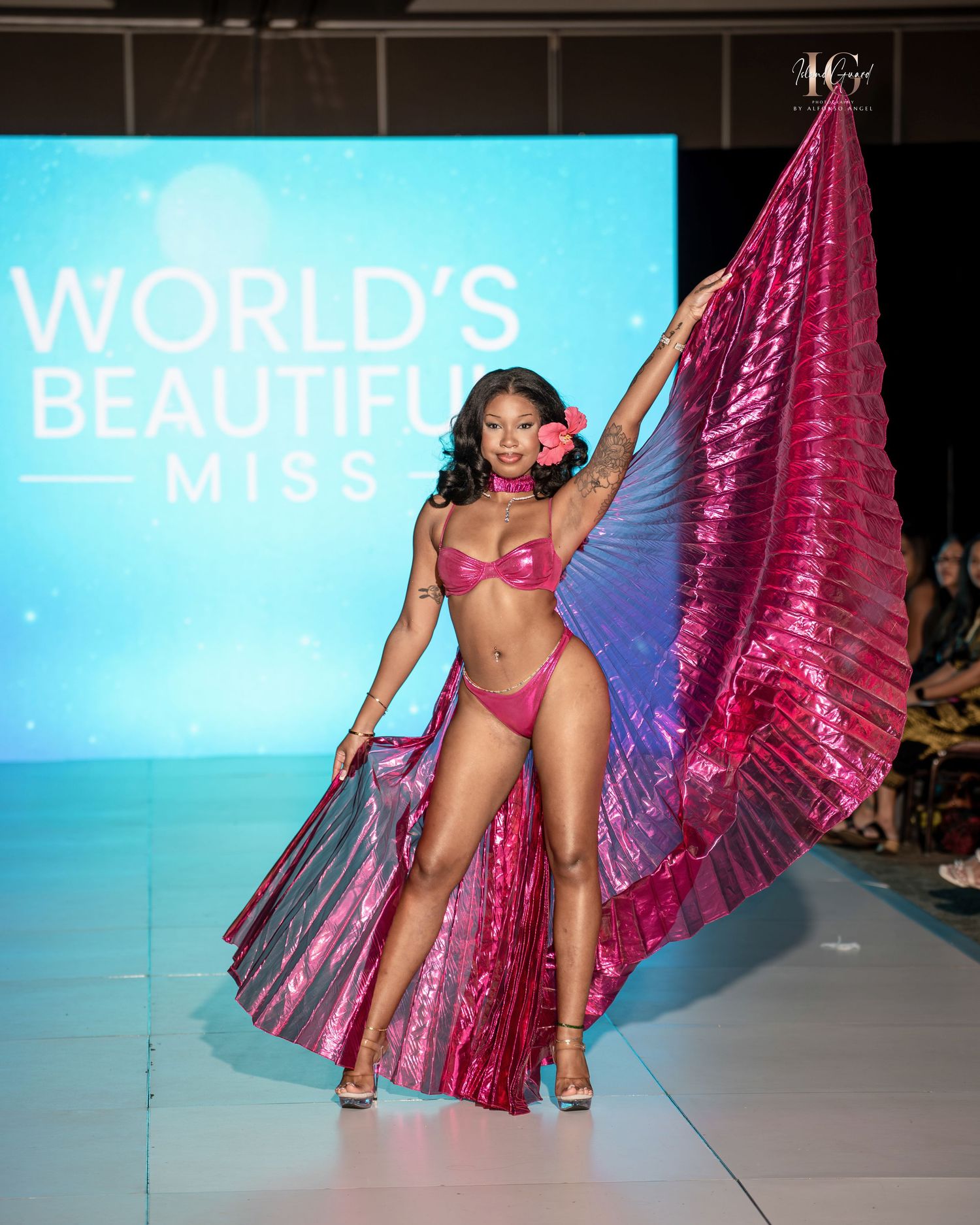Model in pink bikini and flowing magenta cape walks runway at fashion show with blue backdrop.