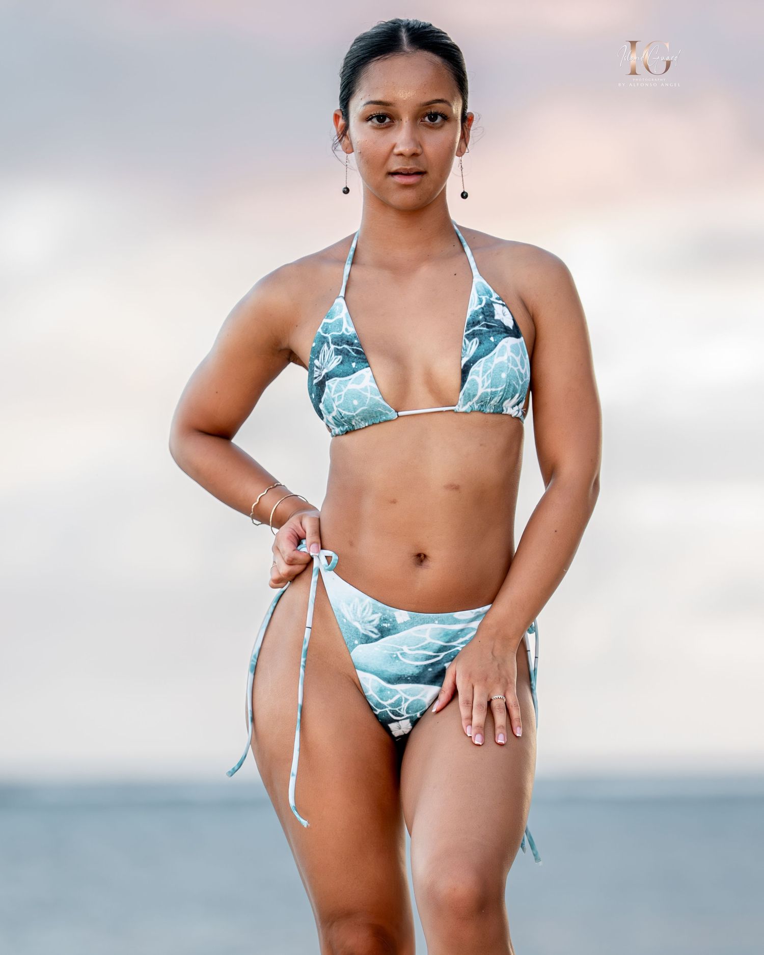 Woman in light blue floral print bikini poses on sandy beach with ocean and cloudy sky in background.