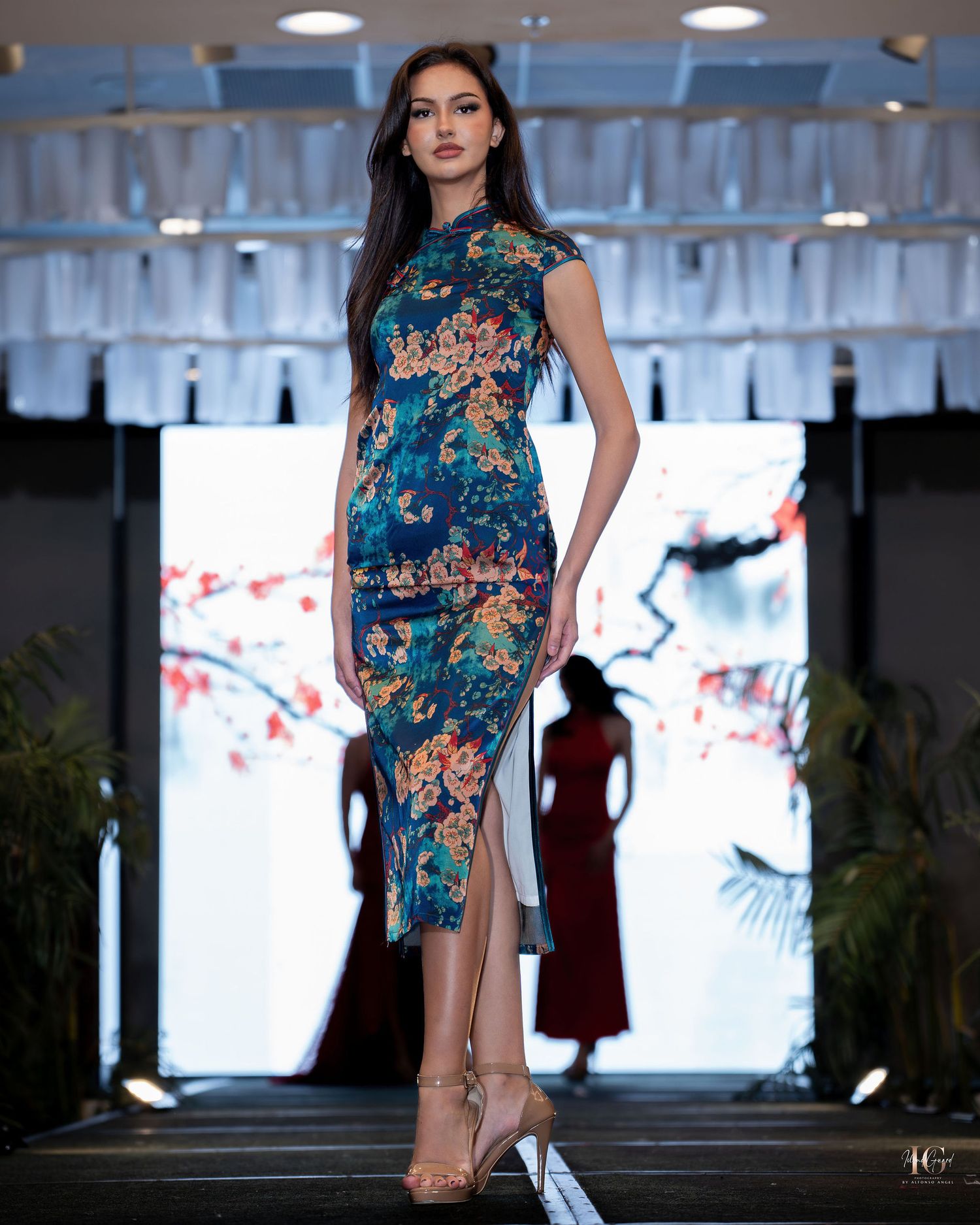 Elegant floral cheongsam-style dress in teal blue with pink blooms shown on runway with cherry blossom backdrop.