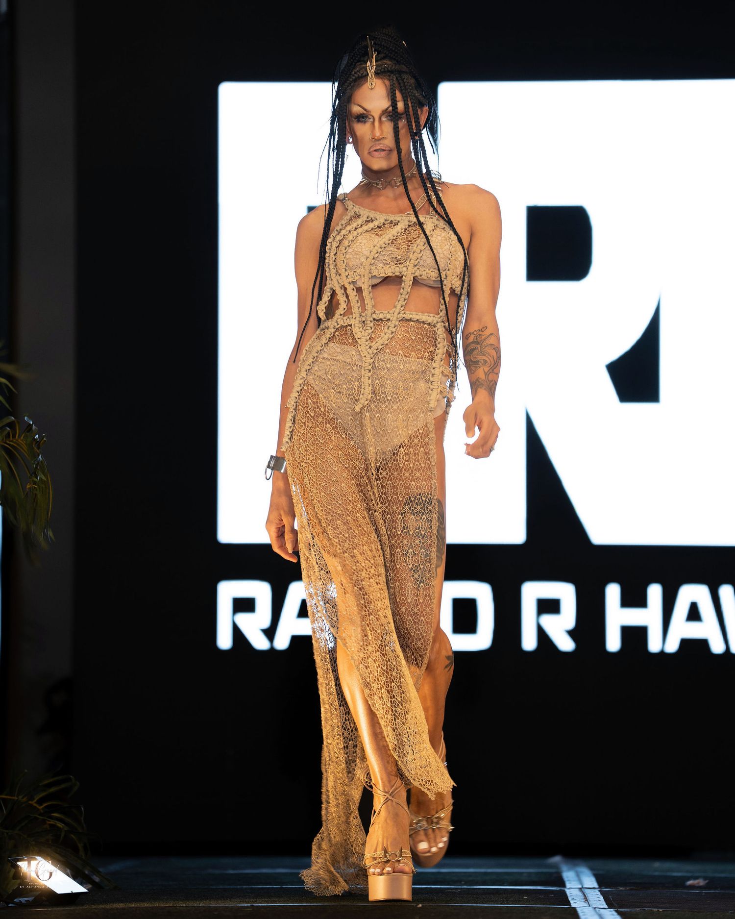 Model walks runway in sheer beige dress with flowing fabric during fashion show against dark backdrop with illuminated text.