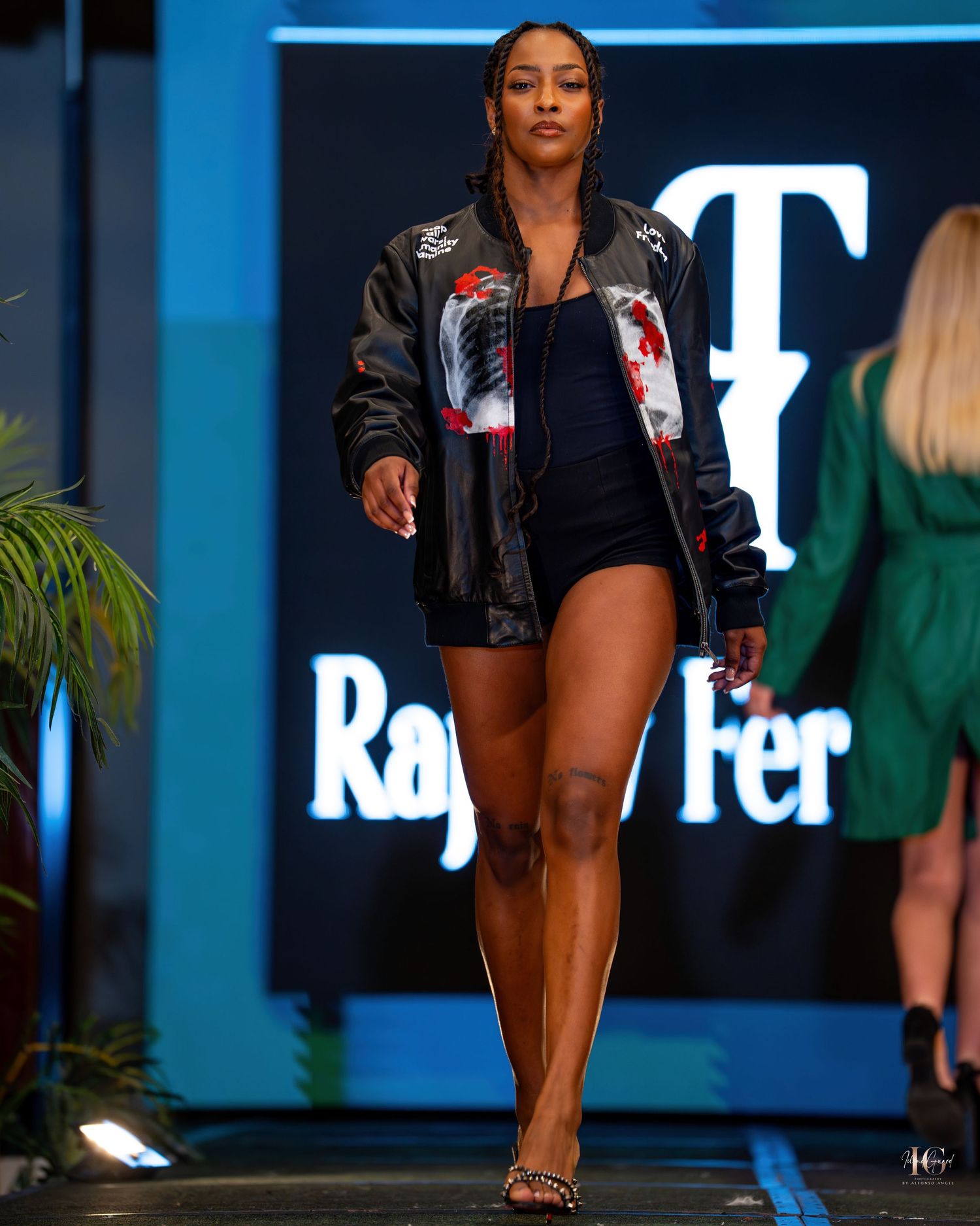Model walks runway in black bodysuit and embellished bomber jacket at fashion show with blue lighting.