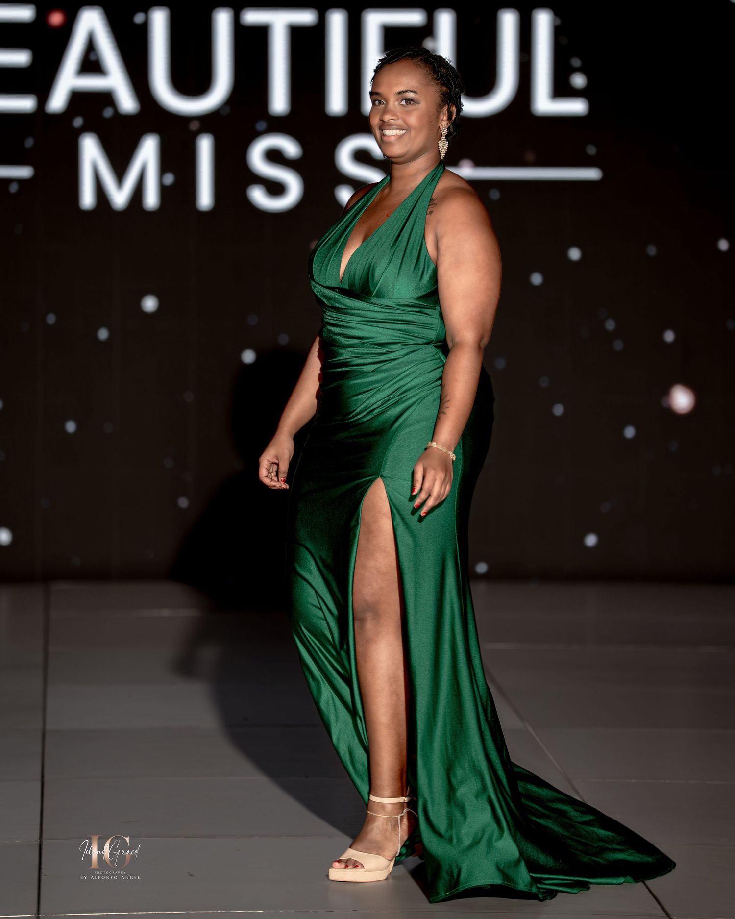 Model walks runway in flowing emerald green evening gown with high slit during fashion show.
