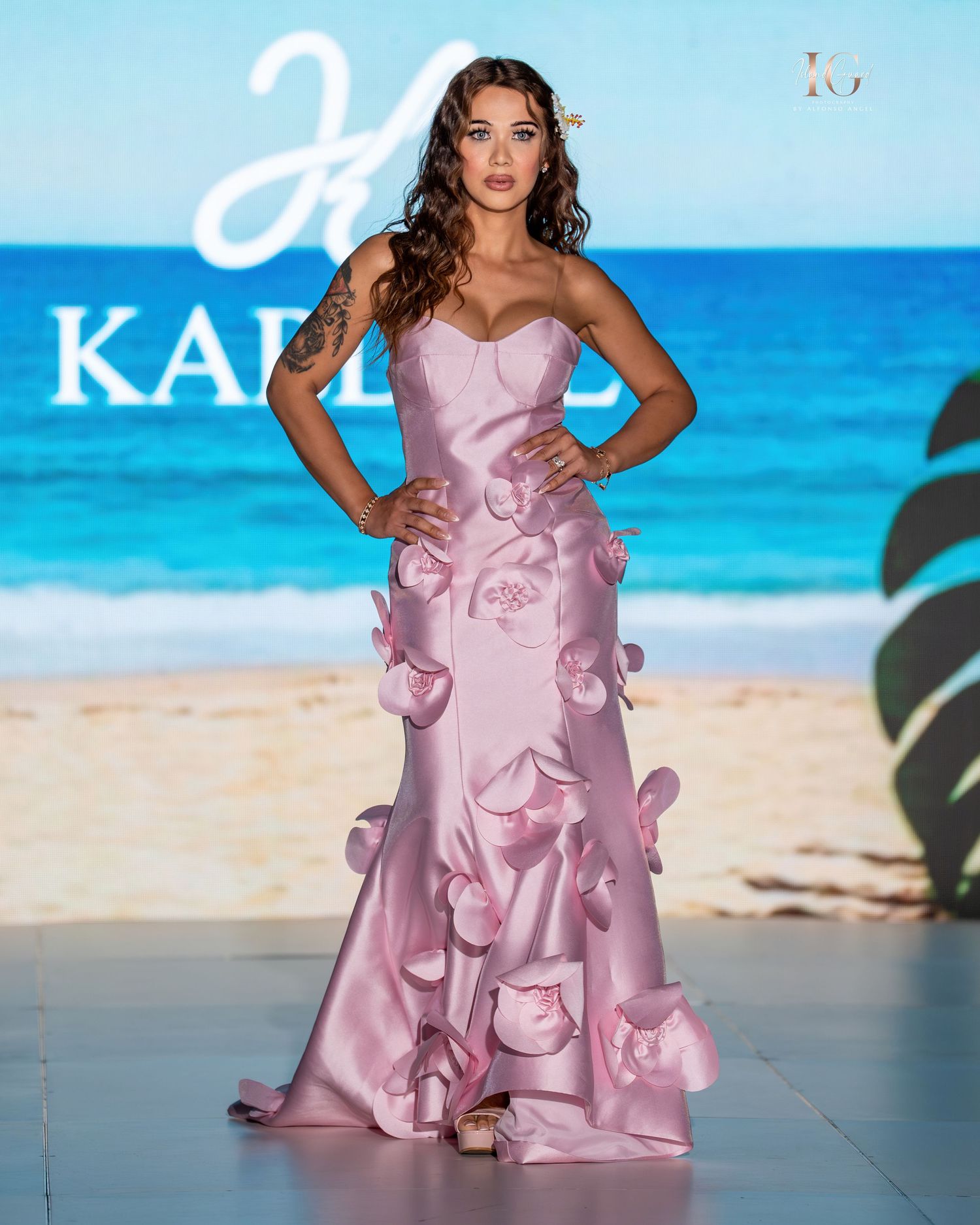 Model walks runway in flowing pink gown with ruffled details against tropical beach backdrop at fashion show.