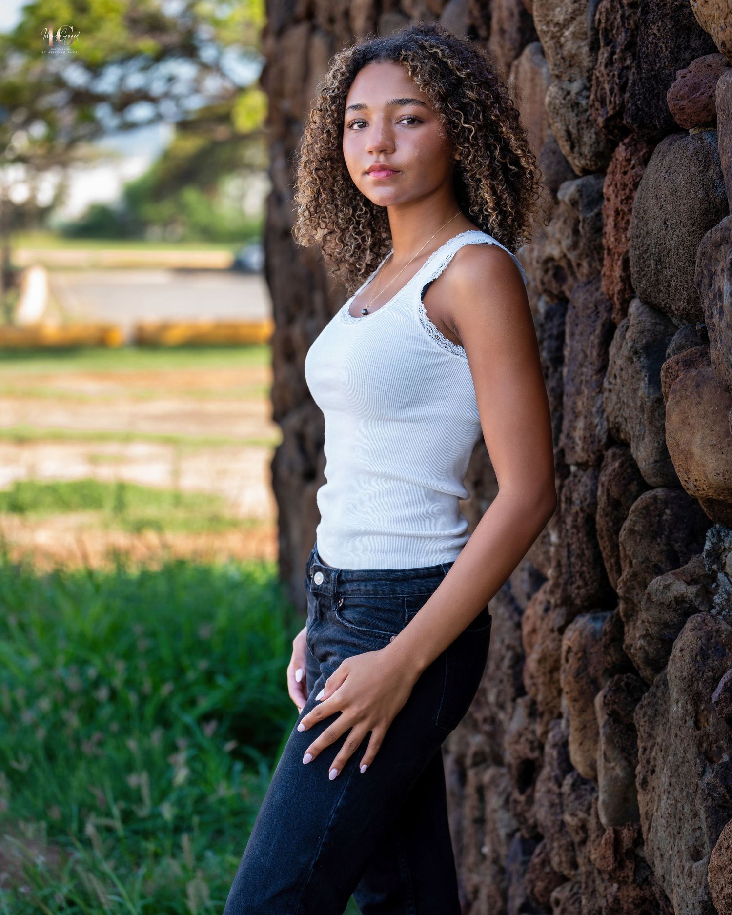Young woman in white tank top poses against rough textured tree bark background in outdoor summer setting.