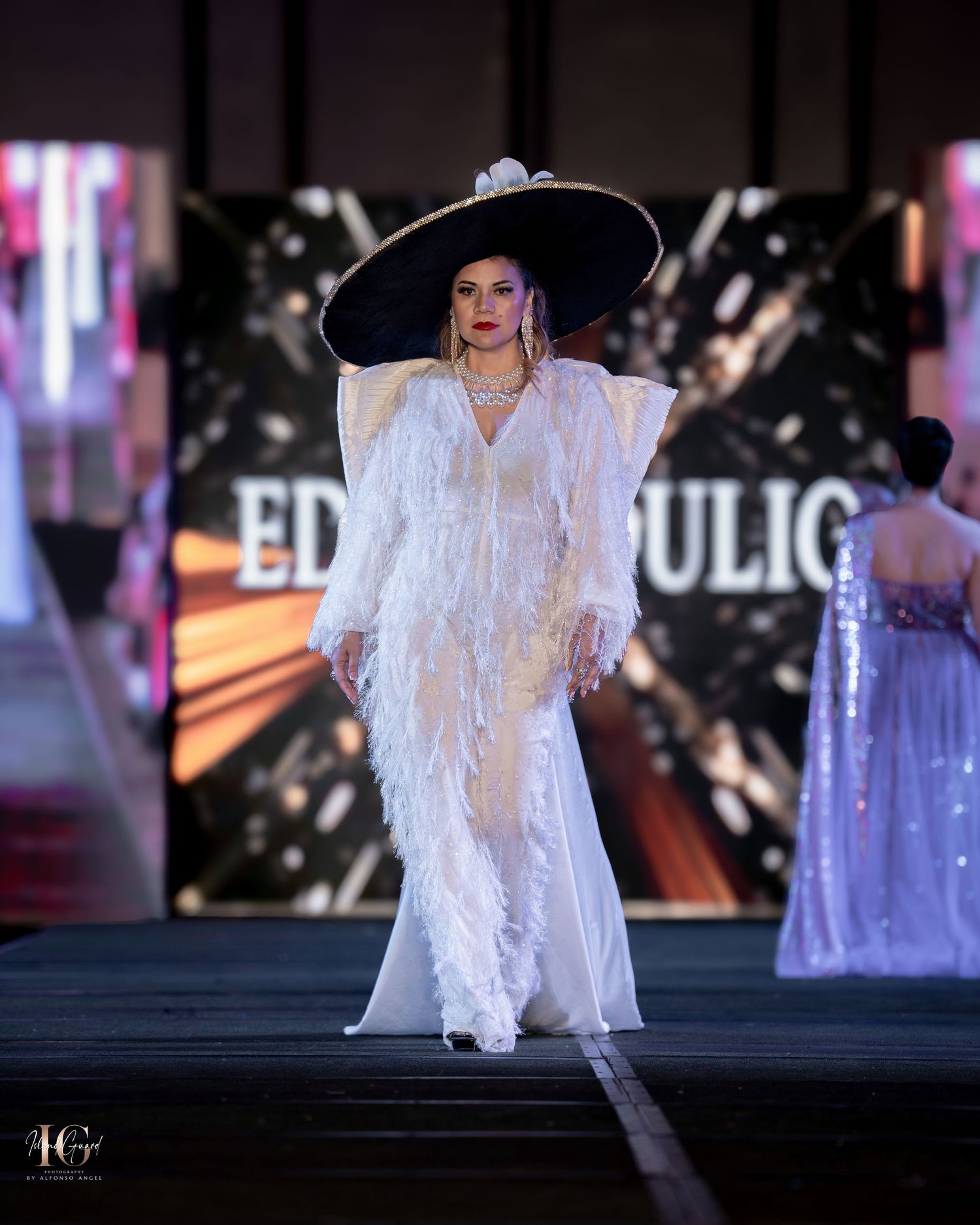 Elegant white lace gown and wide-brimmed hat shown on fashion runway with dramatic lighting.