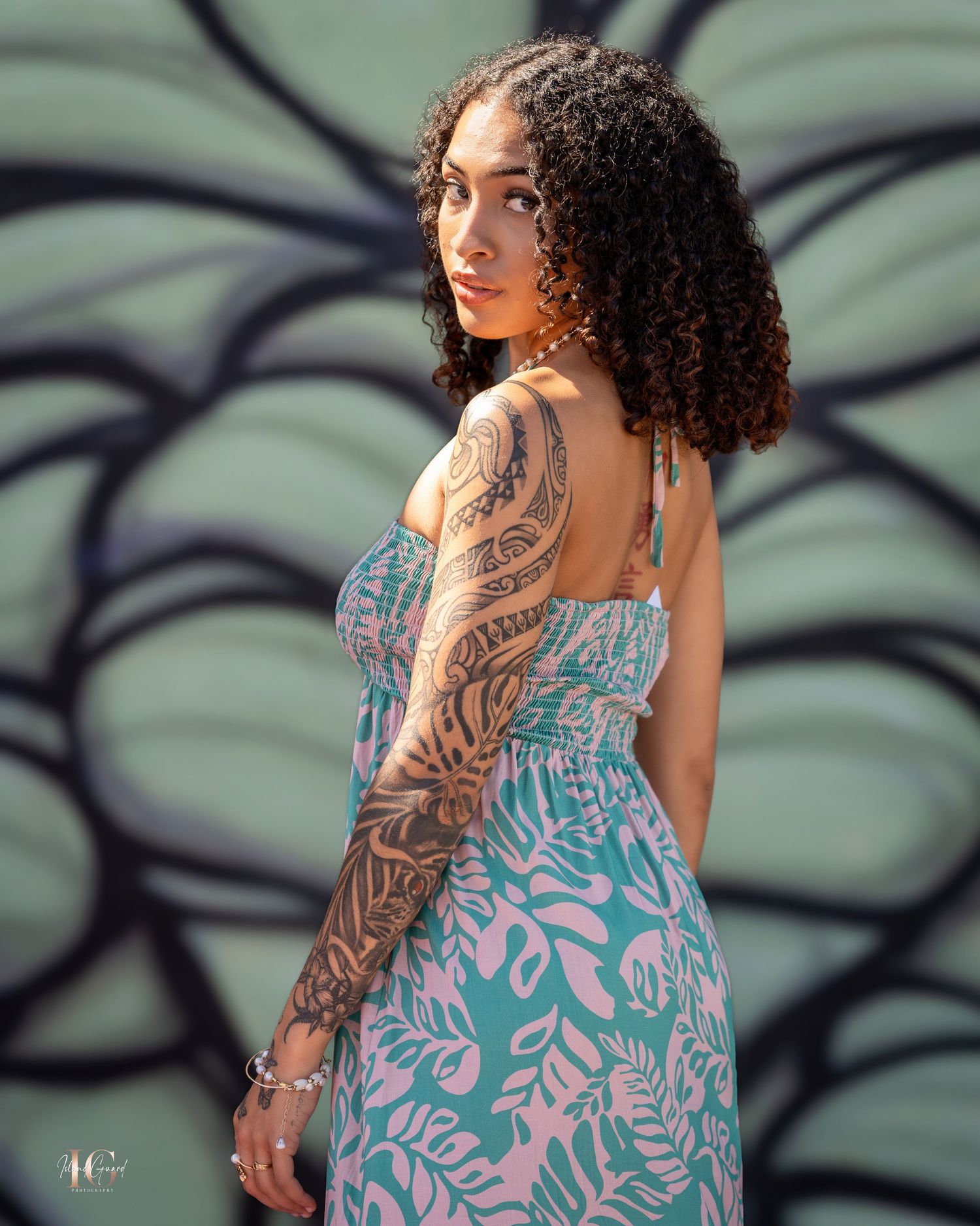 Model in turquoise floral dress shows detailed sleeve tattoo against painted wall background.