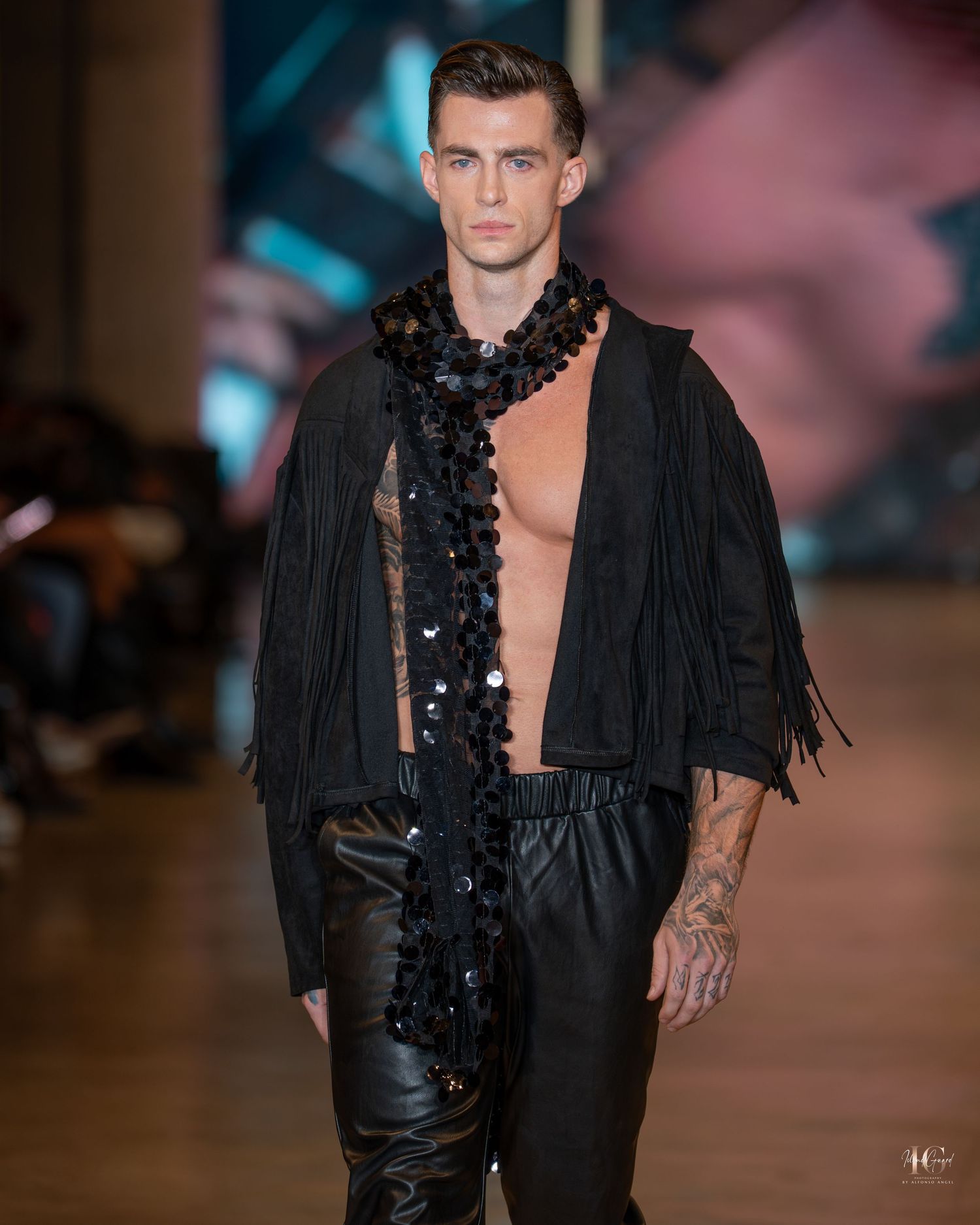 Fashion model walks runway in black leather and fringe outfit with dramatic lace detail at fashion show.