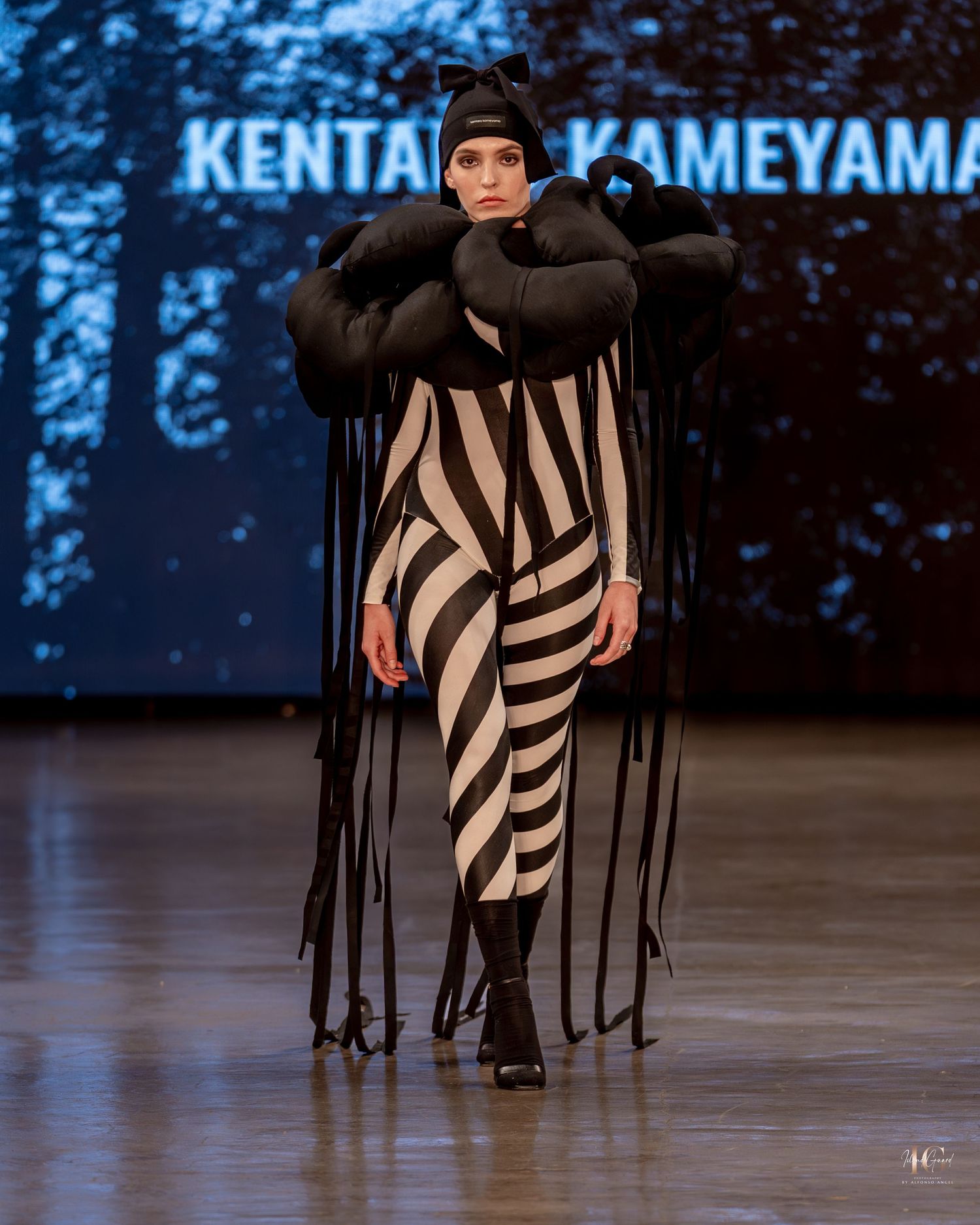 A fashion model walks down a dark runway wearing a dramatic black and white striped outfit with flowing fabric details.