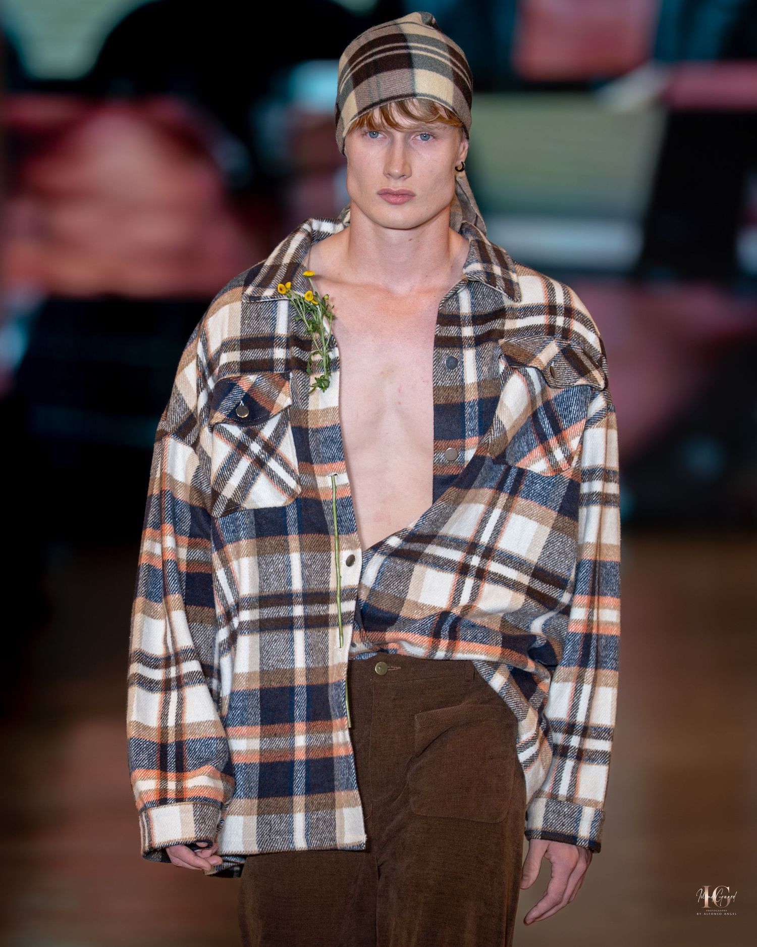 Model walks runway wearing brown plaid flannel shirt and brown corduroy pants at fashion show.