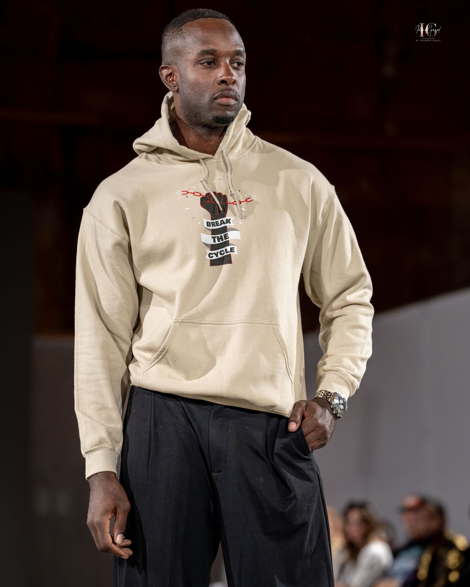 A person wearing a beige hoodie and dark pants poses against a dark background during what appears to be a fashion show.