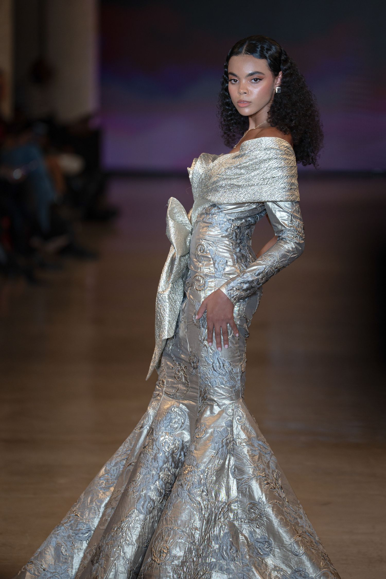 Model walks runway in silver metallic off-shoulder mermaid gown with long sleeves and dramatic bow detail at fashion show.