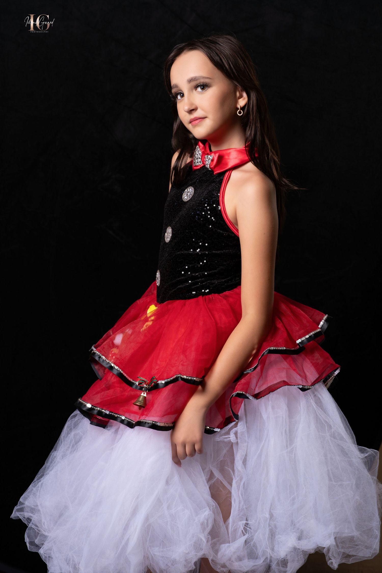 A child wearing a festive red and black tulle dress with white underskirt poses against a dark background for a portrait.