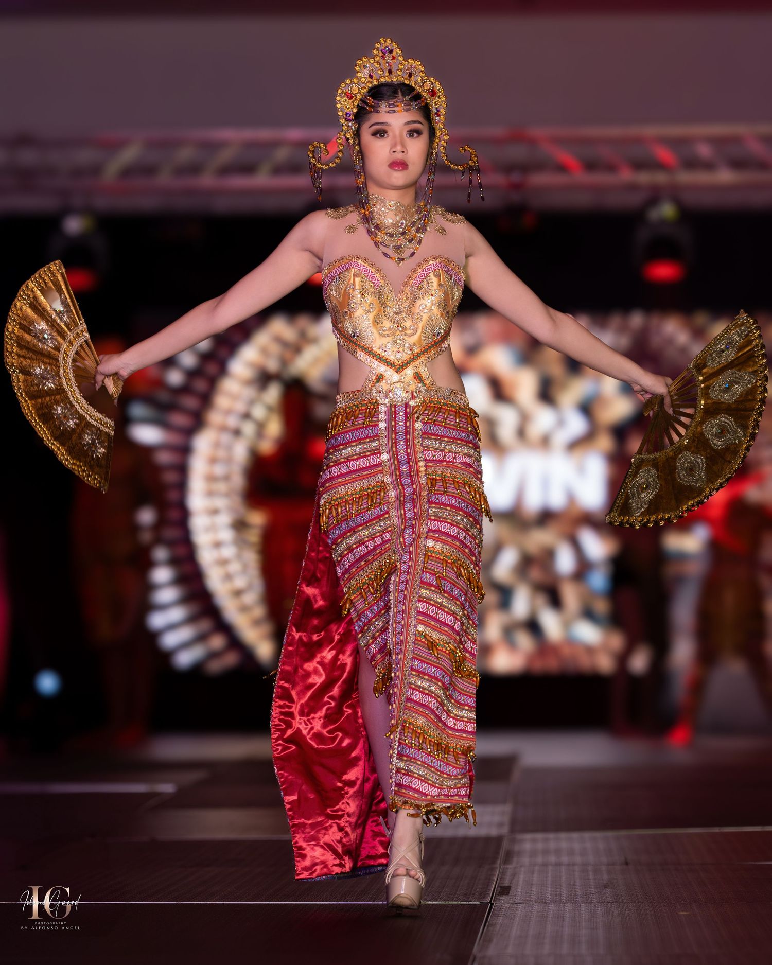 Traditional Thai-inspired golden costume with ornate headdress and decorative fans on fashion runway.
