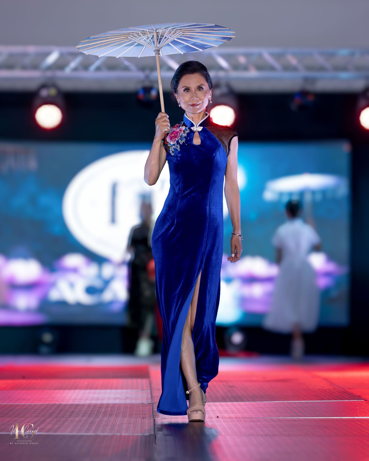 Model in royal blue qipao dress walks runway with parasol at fashion show.
