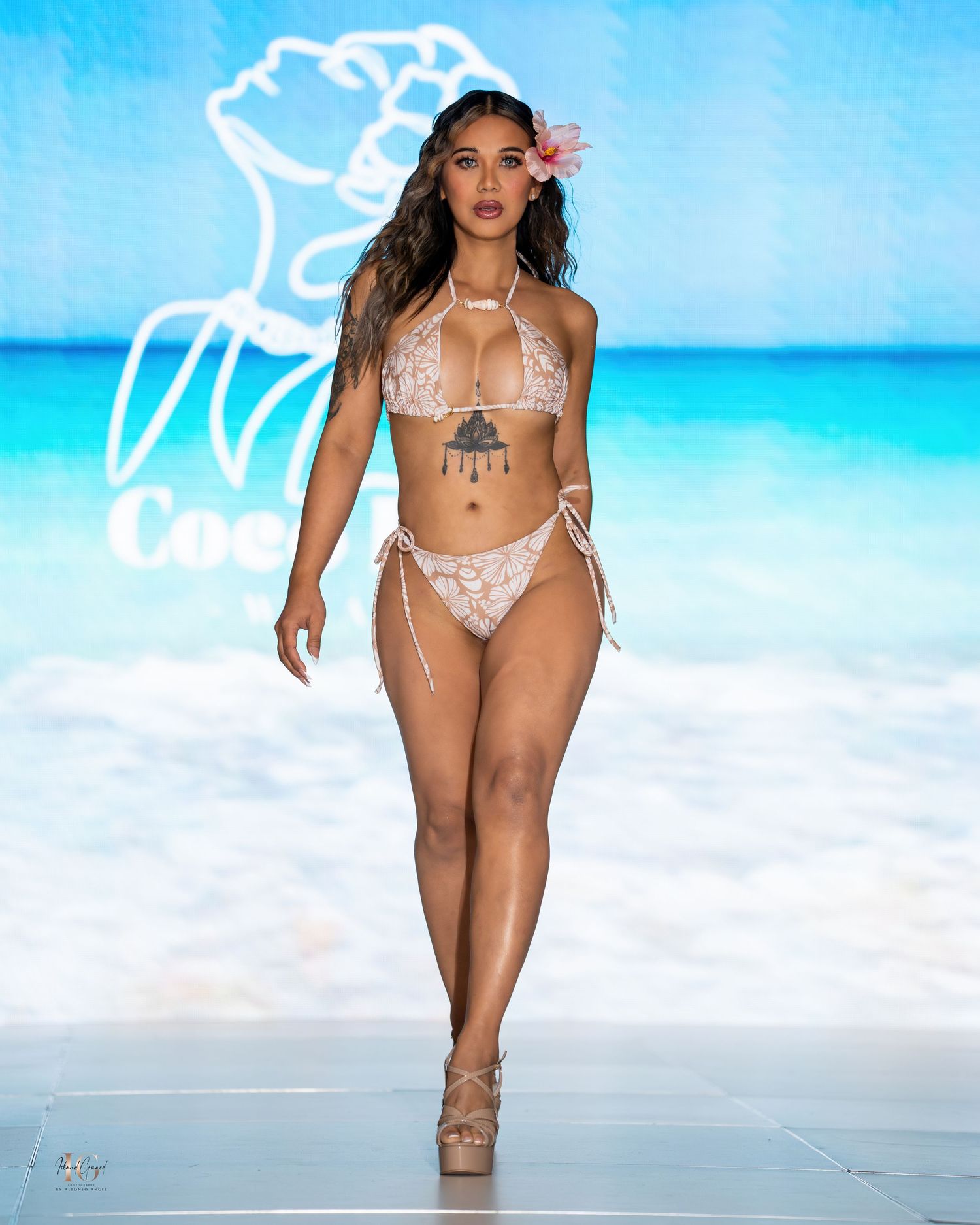 Model showcases delicate beige swimwear with floral accessories at beach-themed fashion show.