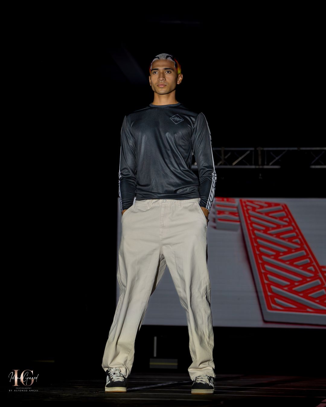 Model wearing black top and khaki pants stands posed on runway with red geometric backdrop.