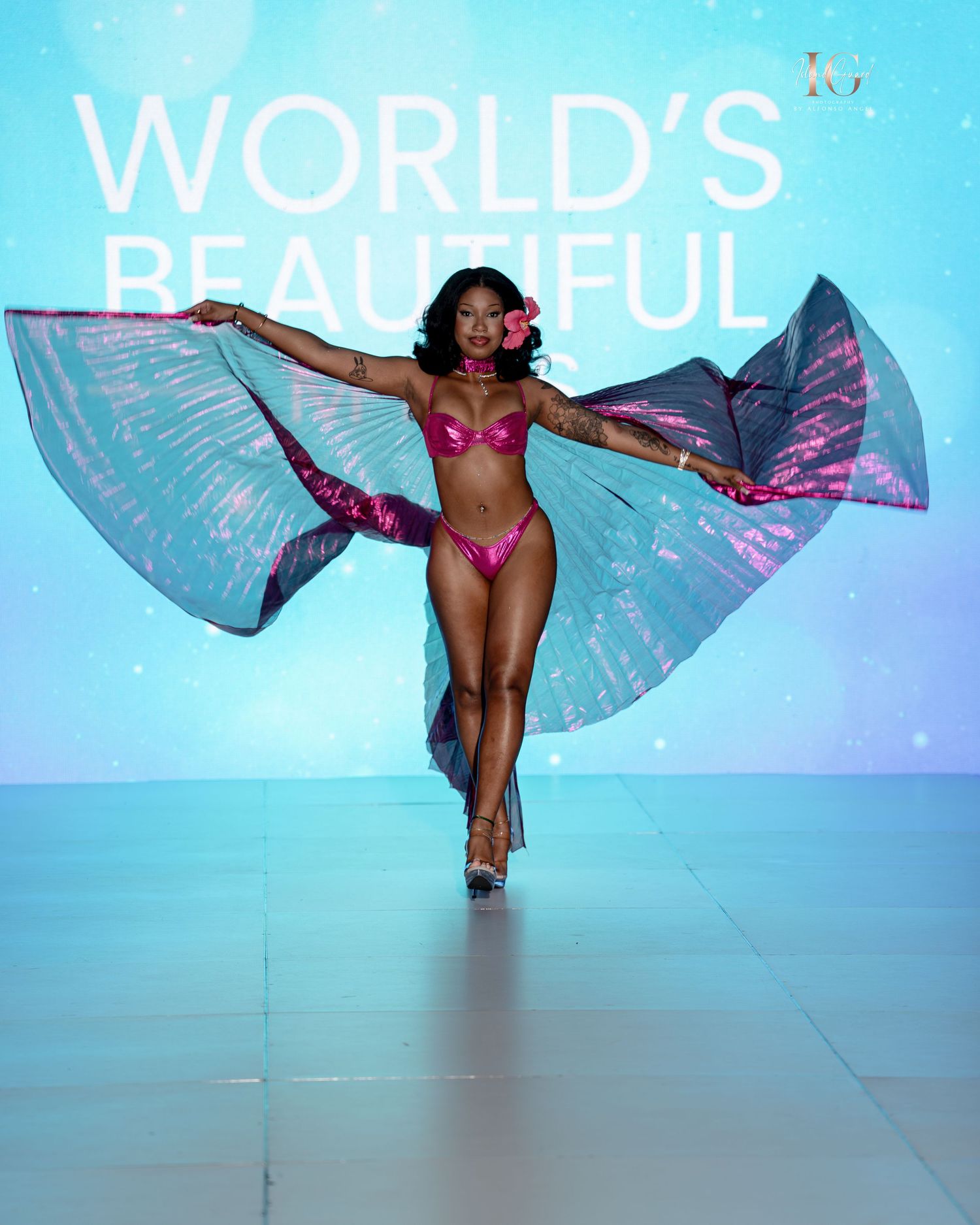 Performer in pink swimsuit dances gracefully with flowing blue and purple fabric wings on a bright illuminated stage.