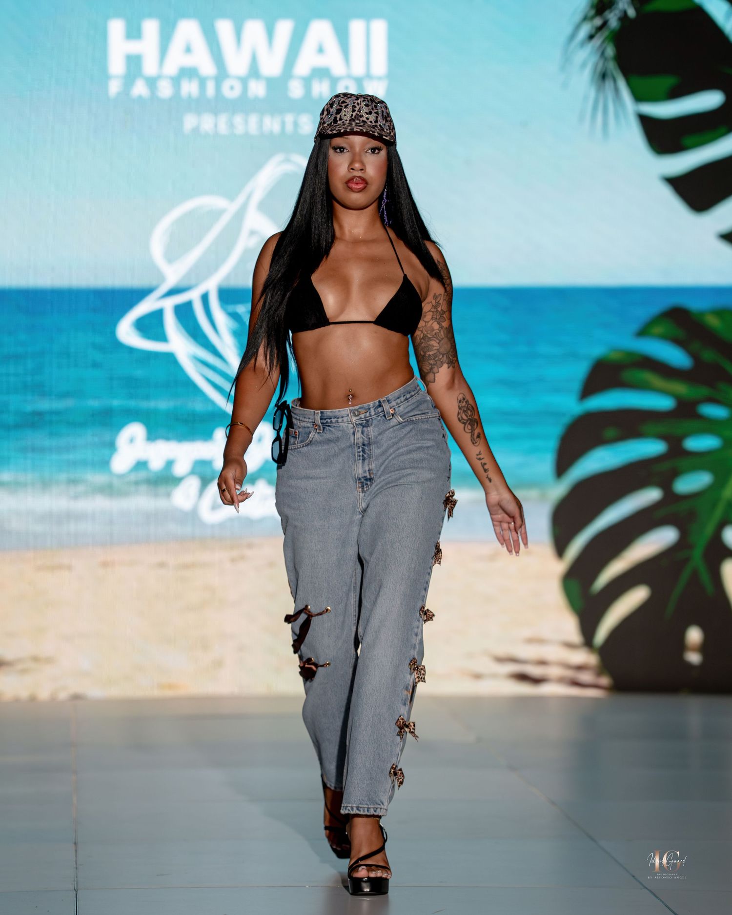 Model walks runway in gray denim ensemble during Hawaii fashion show with tropical beach backdrop.