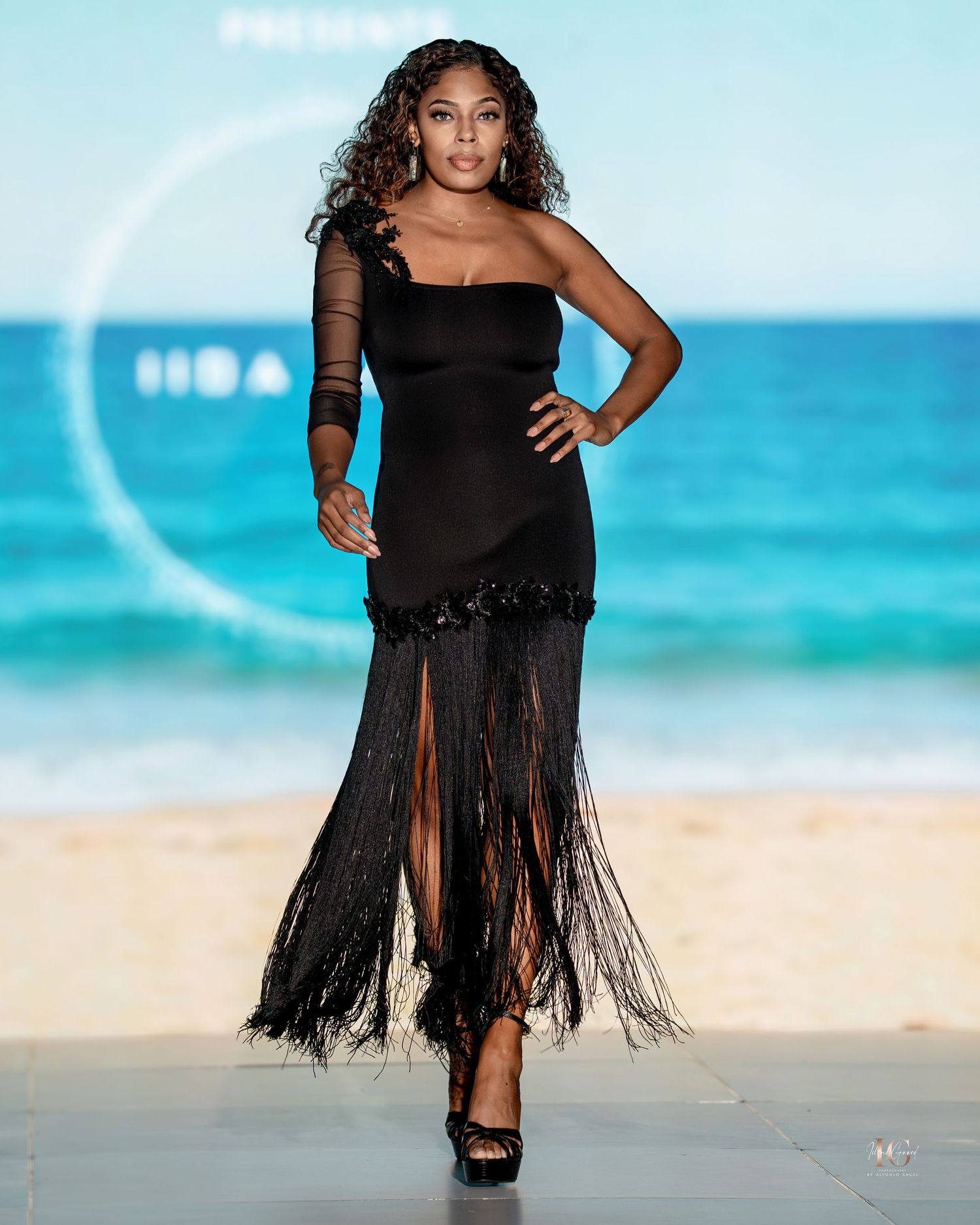 A model in a black fringed dress poses dramatically on a tropical beach with turquoise waters in the background.