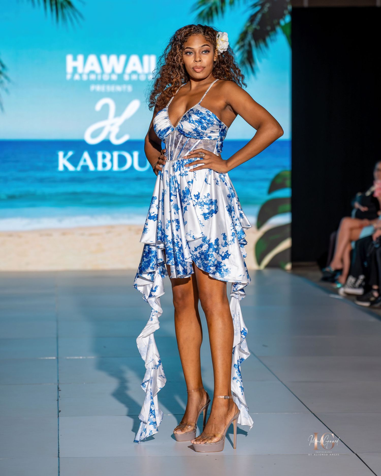 Model walks runway in blue and white tie-dye high-low dress at Hawaii fashion show with beach backdrop.