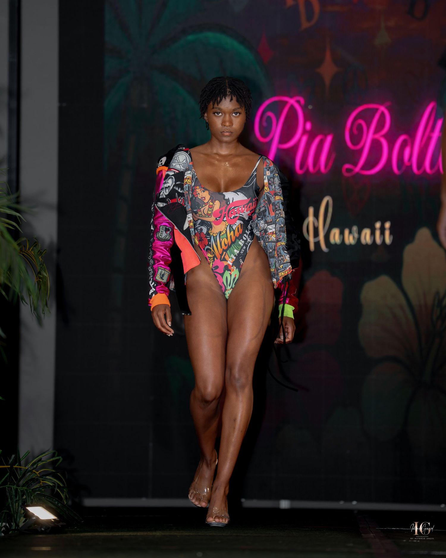 Models walk the runway in colorful swimwear at a Hawaii-themed fashion show with neon pink signage.