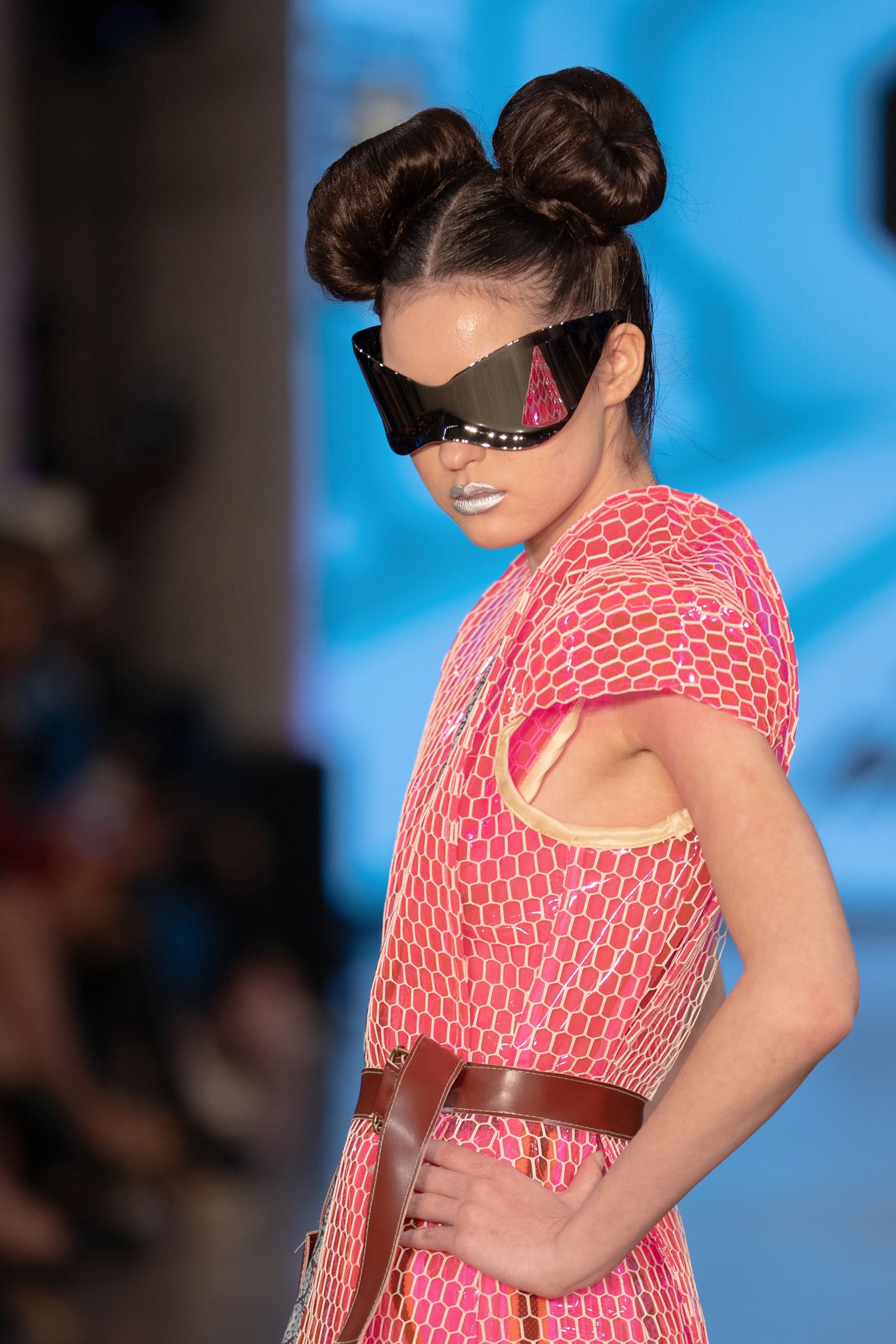 Model walks runway in coral patterned dress with cutout detail against blue backdrop during fashion show.