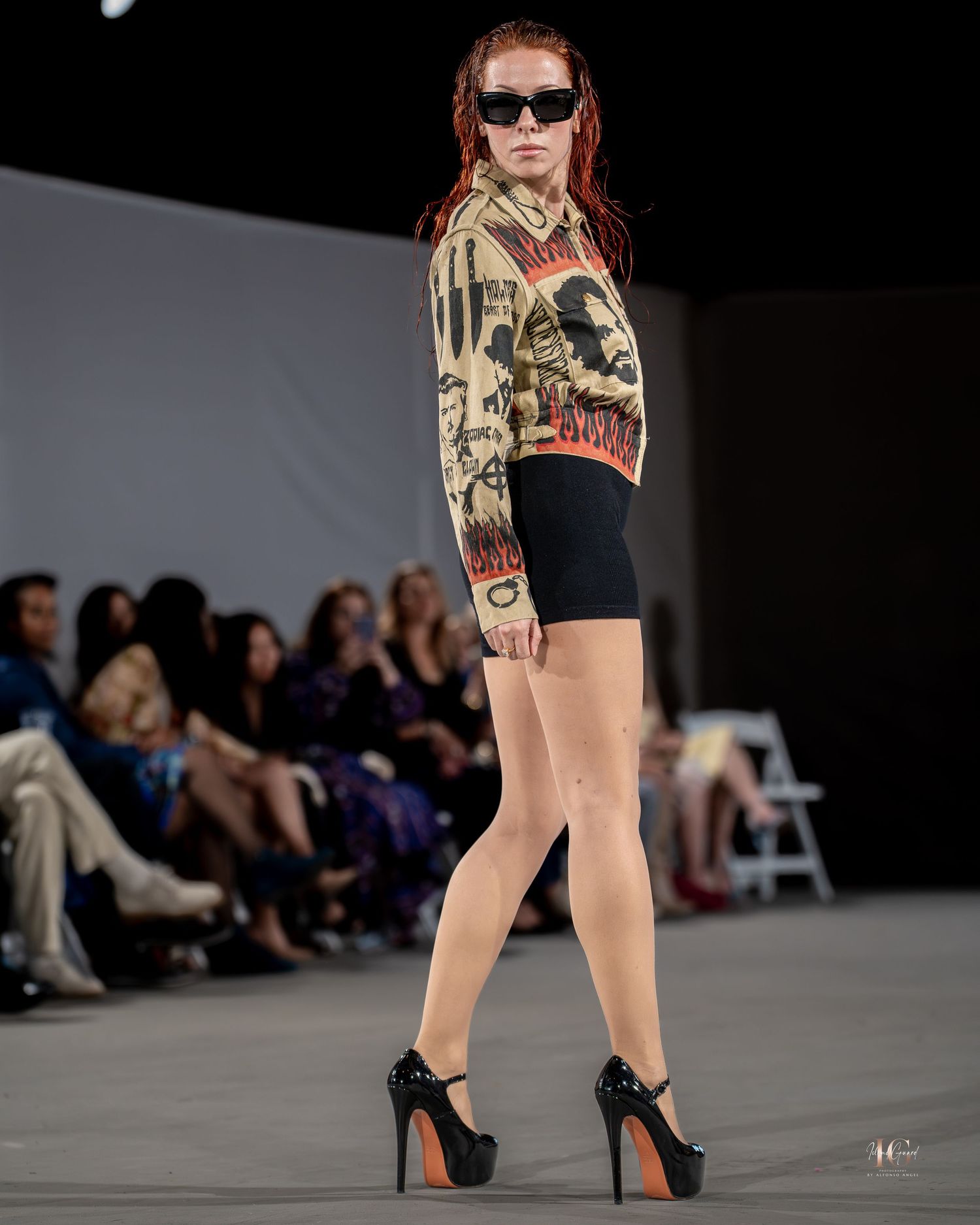 Model walks runway in black heels and printed short dress during fashion show with audience visible in background.