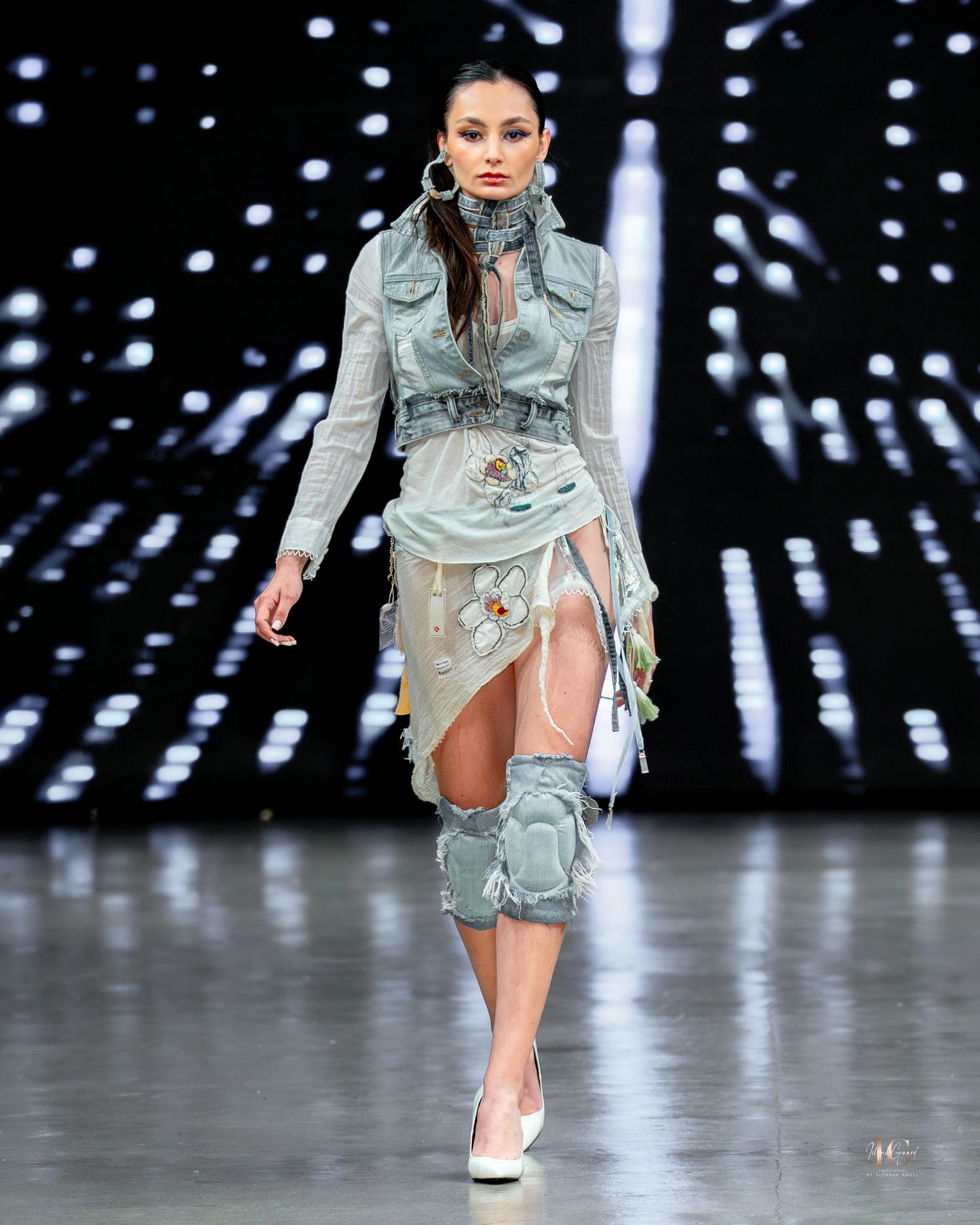 Model walks runway in pale blue distressed military-inspired outfit with knee pads and high collar during fashion show.