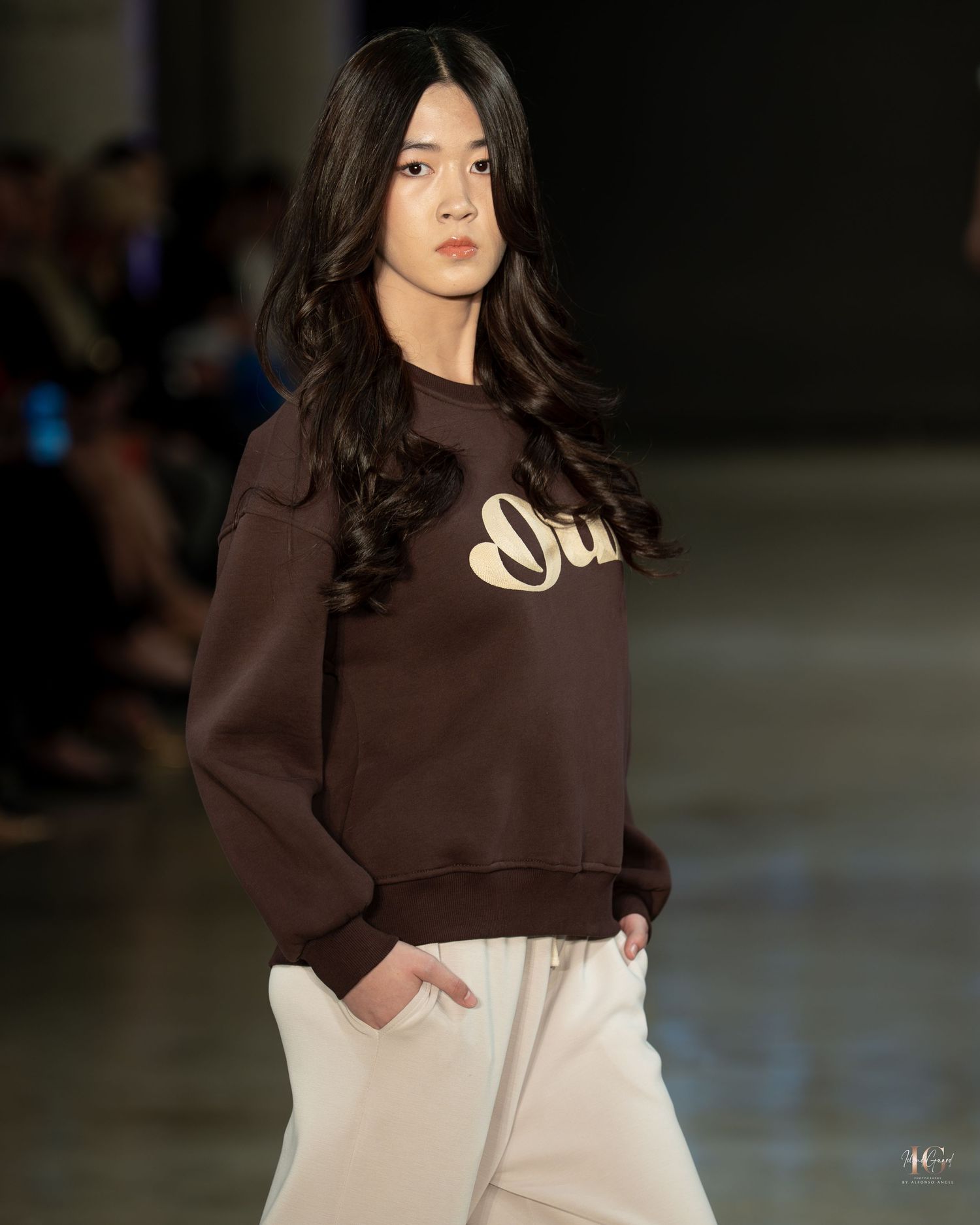 Model walks runway wearing brown sweatshirt with text and white pants during fashion show.
