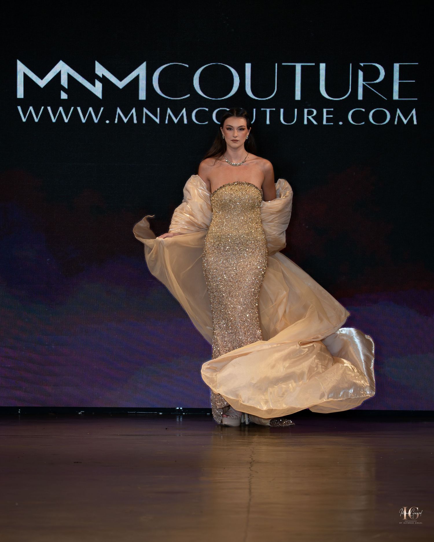 A model walks the runway in a flowing champagne-colored gown with a dramatic twirl at a fashion show.