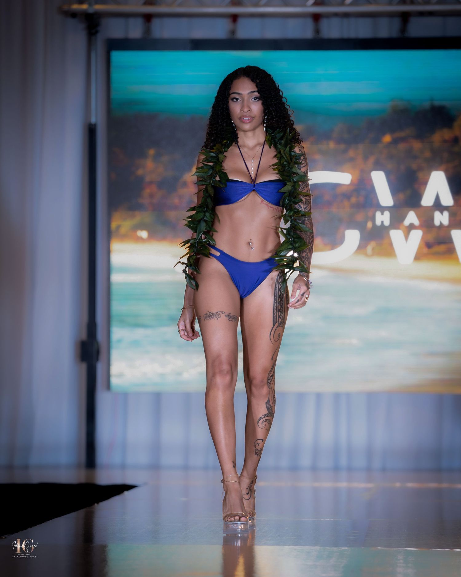 Swimwear models showcase blue bikinis on an illuminated catwalk with beach backdrop.