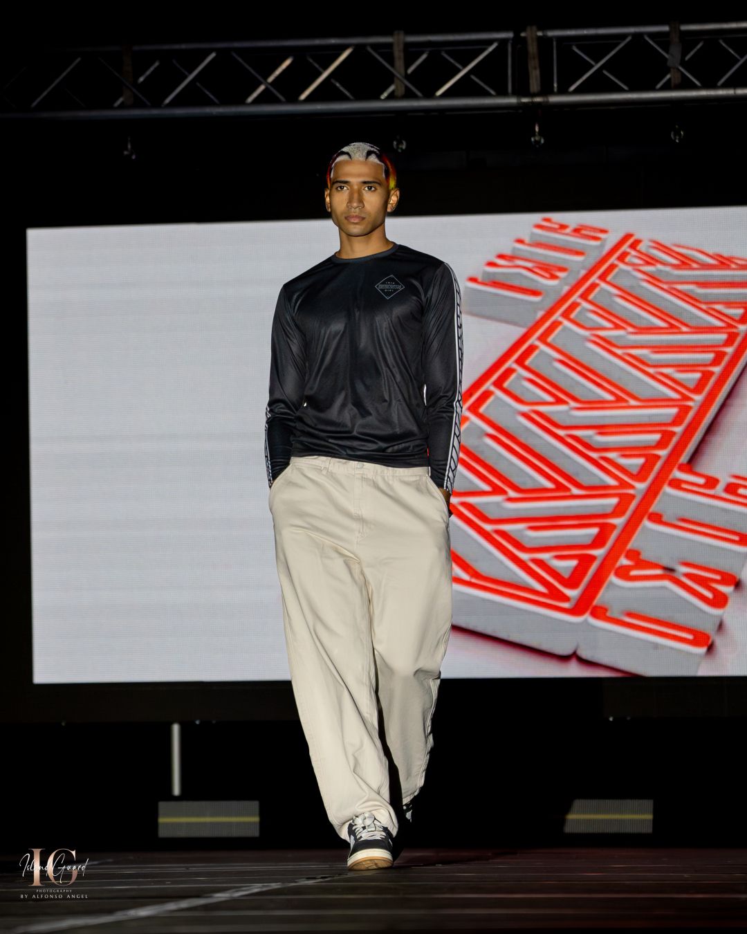 Model in black long sleeve top and beige pants walks runway with geometric red backdrop.