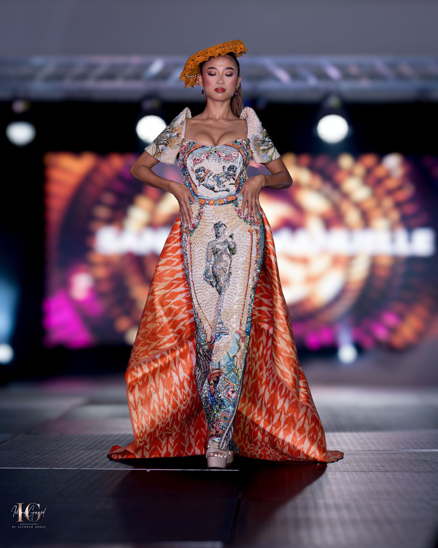 Haute couture gown with silver bodice and dramatic orange cape strikes pose on fashion runway.